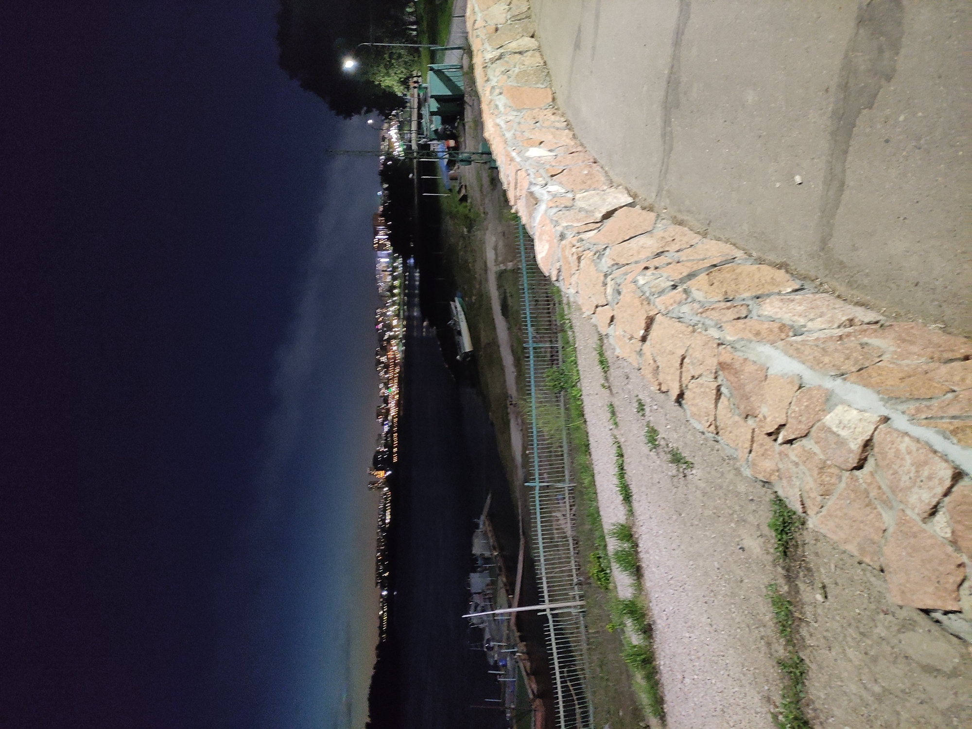 The image depicts a night scene along the Irtysh River in Pavlodar, Kazakhstan. The foreground features a stone embankment, partially illuminated, with a paved walkway above it. A metal fence runs along the edge of the embankment, separating it from the river.
In the middle ground, the dark waters of the Irtysh River are visible, with several boats or small vessels moored along the bank. On the opposite shore, a cityscape is illuminated with numerous lights, indicating an urban area.
The background shows a dark night sky, transitioning to a lighter hue above the city lights. Streetlights are visible along the embankment and in the distance, contributing to the overall illumination of the scene. The image appears to be taken from a high vantage point, providing a wide view of the river and cityscape.
