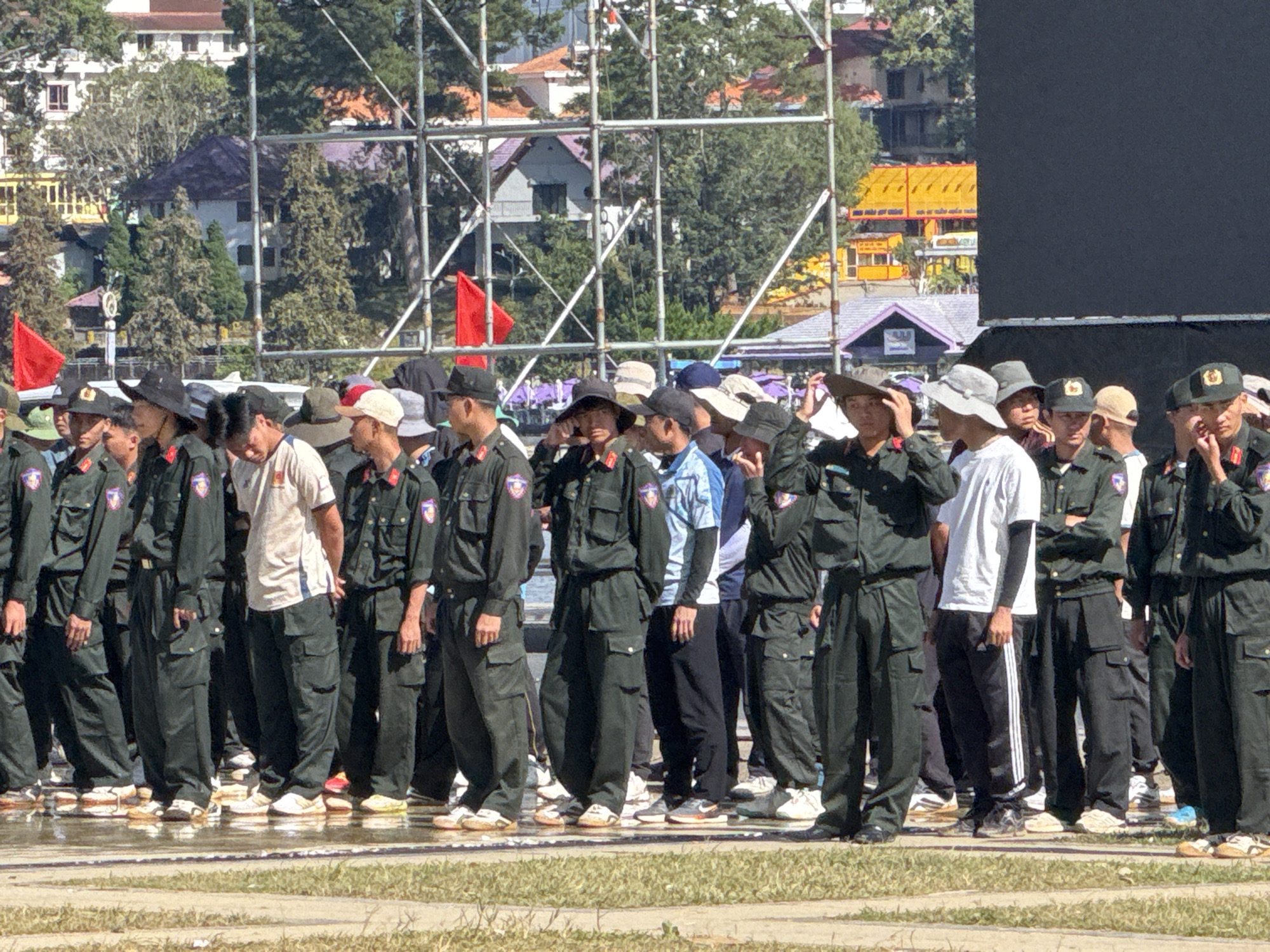 The image depicts a group of individuals, most of whom are wearing dark green military-style uniforms with emblems on their sleeves. They are standing in a line outdoors on what appears to be a paved or reflective surface, possibly a stage or promenade. Many of the individuals are wearing hats, including some wide-brimmed hats and others that are more fitted. Some are looking forward, while others appear to be conversing or observing their surroundings.
In the background, there are buildings with distinctive architecture, suggesting an urban or semi-urban setting. Some of the buildings have red-tiled roofs and multiple stories. There are also many trees, indicating a location with significant greenery. A metal scaffolding structure is visible behind the group, and a large black screen or banner is positioned to the right of the frame. Red flags are also present, adding a splash of color to the scene. The overall impression is of an organized gathering, possibly a ceremony or event.
Based on the architectural style of the buildings, particularly the roofs and the presence of lush greenery and hilly terrain, this scene is likely located in Da Lat, Vietnam, a city known for its French colonial architecture and cooler climate. The presence of military-style uniforms could suggest a civic or official event.