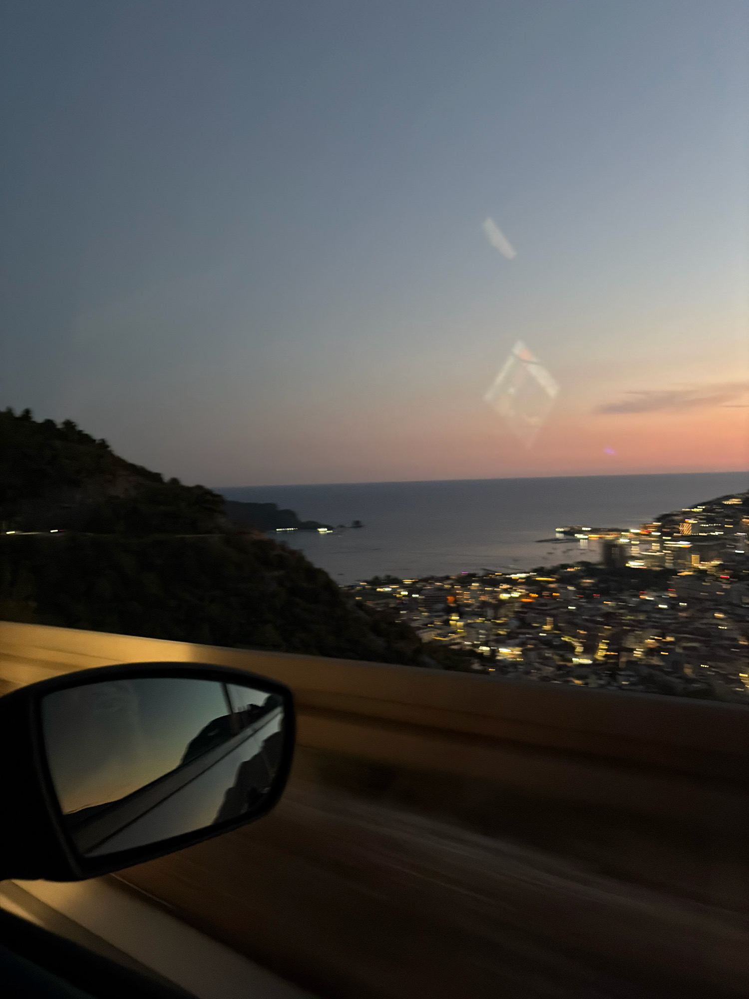 The image captures a view from a vehicle traveling along a coastal road at dusk, overlooking the city of Budva, Montenegro. In the foreground, a side-view mirror reflects the road and the sky. Beyond the mirror, the dark, tree-covered slopes of a mountain descend towards the sea. The distant water is a deep gray, meeting the horizon where a soft, peachy sunset is visible. The city of Budva is illuminated with a multitude of bright lights, giving it a warm glow against the darkening landscape. Buildings are densely packed along the coastline and up the hillsides, suggesting a vibrant urban area. The overall impression is one of a scenic drive at twilight, with the lights of the city emerging as night begins to fall.