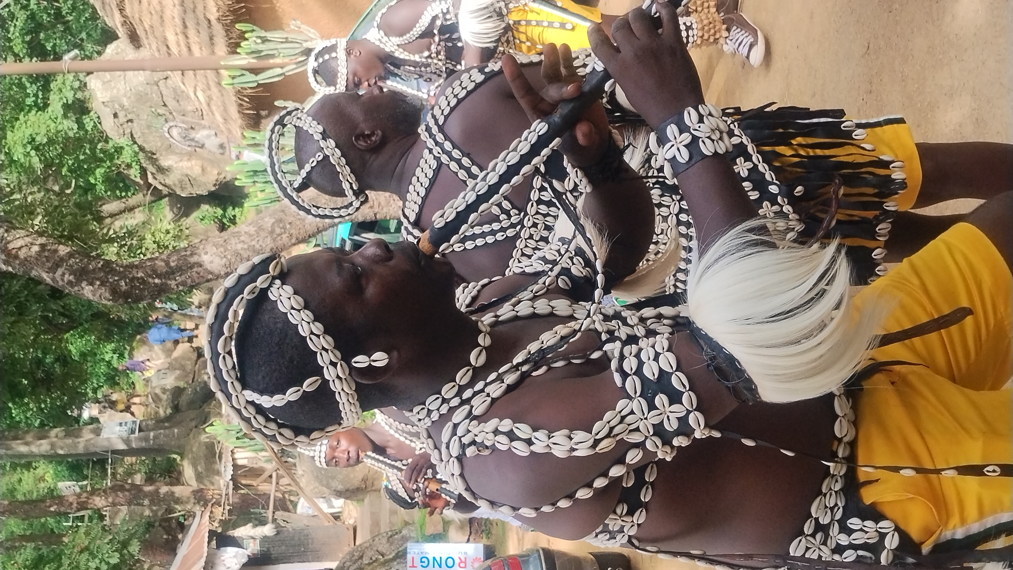 In the city of Jos, Nigeria, a group of men in traditional attire, adorned with cowrie shells, are engaged in a cultural performance. The primary focus is on a man in the foreground, playing a dark, wooden flute. He wears a headdress made of cowrie shells and black material, a necklace of multiple strands of cowrie shells, and armbands also decorated with shells. His outfit includes a black garment with fringed yellow accents, and a large bunch of white hair attached to his left wrist. To his right, another man, also wearing a similar cowrie shell headdress and adornments, is partially visible. In the background, other individuals in similar attire are seen, some also holding flutes. The setting appears to be an outdoor gathering, possibly a festival or ceremony, with trees, rocks, and traditional thatched-roof structures indicating a rural or semi-rural environment. Some people are seen walking up a rocky incline in the distance.