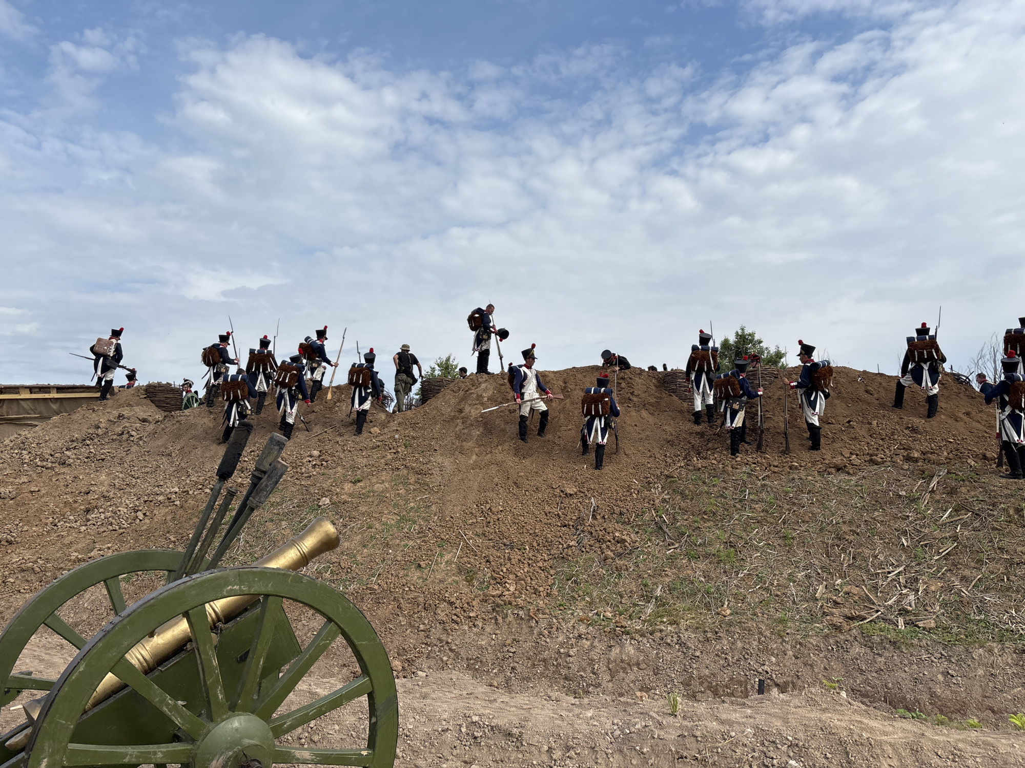 A historical reenactment of a battle is taking place in Goncharovo, Russia. The scene is set on a dirt hill with a cloudy sky overhead. In the foreground, a green cannon with a golden barrel is positioned. A group of soldiers in historical military uniforms, predominantly blue and white with red accents and tall hats, are scattered across the hillside. Some are holding muskets, while others appear to be moving or preparing for action. The soldiers are diverse in their positions, with some higher up on the slope and others lower down. The environment is natural, with dirt, grass, and scattered dry vegetation. The overall impression is a staged historical event, possibly a reenactment of a Napoleonic-era battle given the uniforms.