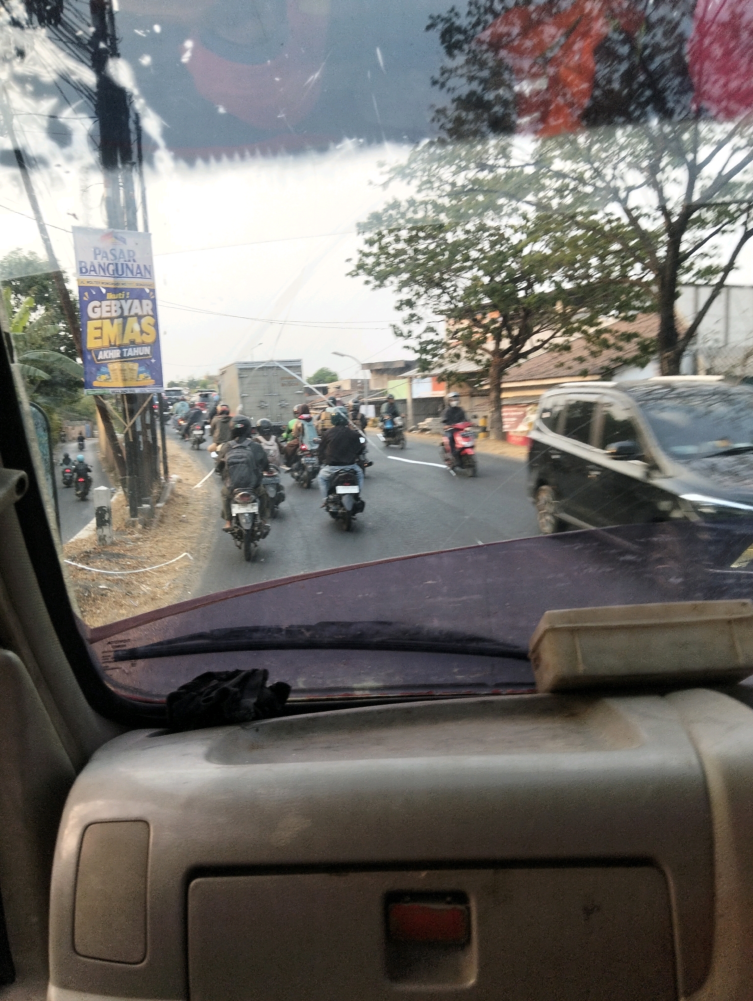 The image is a point-of-view shot from inside a vehicle looking through the windshield. The scene depicts a busy street in Semarang, Indonesia, characterized by a large number of motorcycles in traffic. A prominent blue and yellow billboard with Indonesian text, "PASAR BANGUNAN GEBYAR EMAS AKHIR TAHUN," is visible on the left side of the road. The traffic includes many motorcycles, some with riders wearing helmets, and a few cars, including a dark-colored SUV on the right. Trees and buildings line the street. The interior of the vehicle shows the dashboard with a small box-like object on it and a black glove placed on the dashboard. The overall lighting suggests it is daytime, possibly late afternoon, given the shadows and the bright sky.