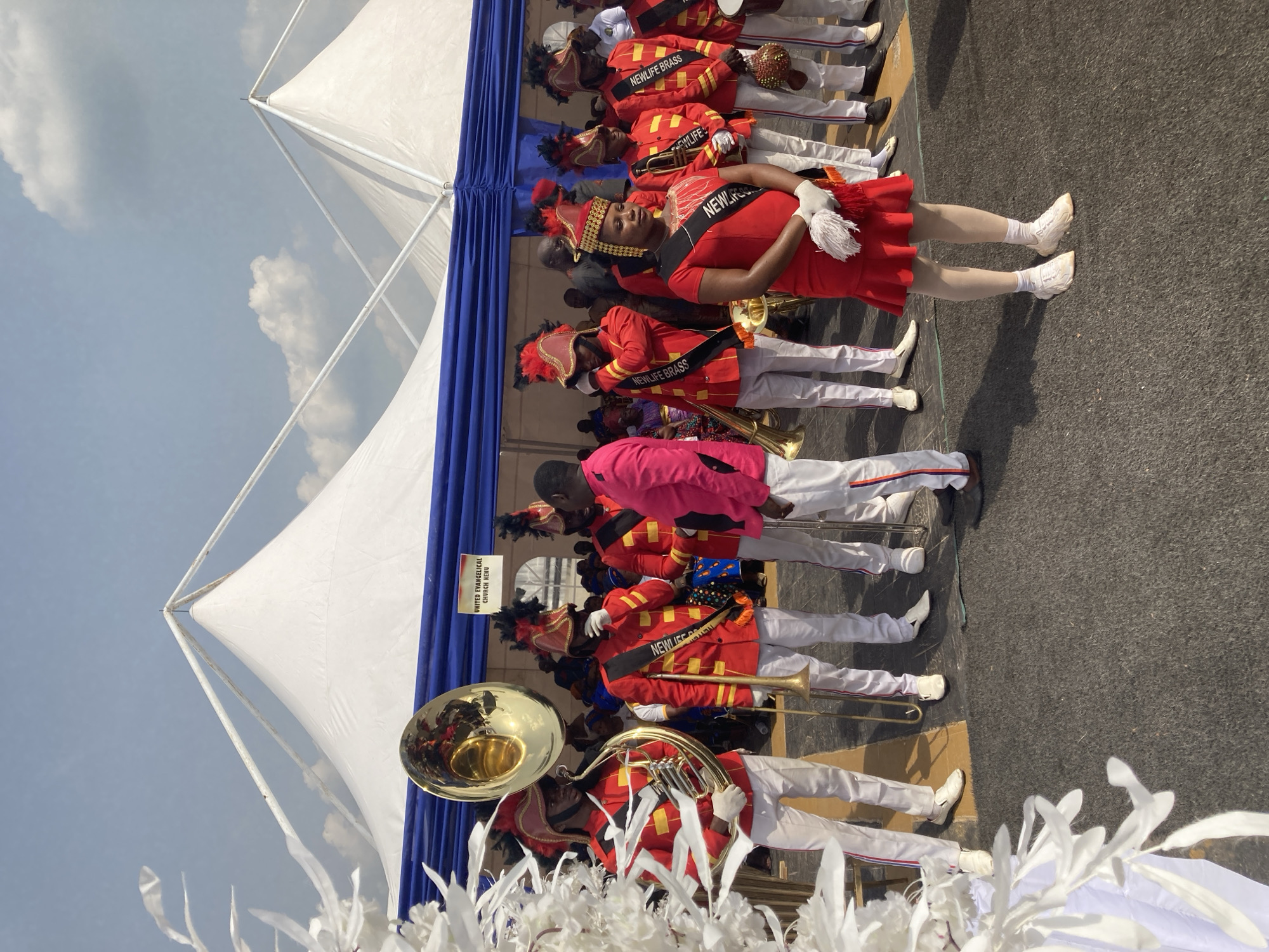 A New Life Brass band is performing outdoors in Nnenu, Nigeria under a white tent with blue draping. The band members are wearing red jackets with gold trim, white pants, and ornate hats. The woman in the front is wearing a red dress and a sash that reads "New Life Brass." The band members are holding brass instruments. There is a sign that reads "United Evangelical Church, Nnenu." White feathered decorations are in the foreground.