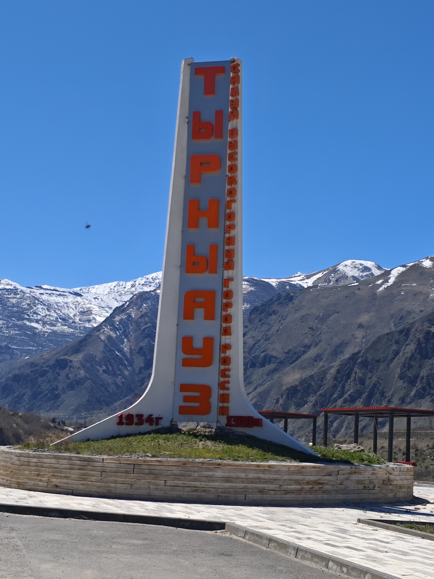 A tall, gray monument with red Cyrillic text stands prominently in the foreground. The text spells out "Tyrnyauz". The base of the monument is adorned with the year "1934" in red. The monument is situated on a raised circular platform with a retaining wall made of light-colored stone or concrete blocks. Short grass and some weeds grow on the platform.
The background features a mountainous landscape with snow-capped peaks. The sky is clear and bright blue. A dark object, possibly a bird or insect, is visible in the sky.
In the mid-ground, a structure with a red roof and dark support beams can be seen behind the monument, suggesting some sort of pavilion or sheltered area. The overall scene is a daytime shot of a city welcome sign located in a mountainous region.