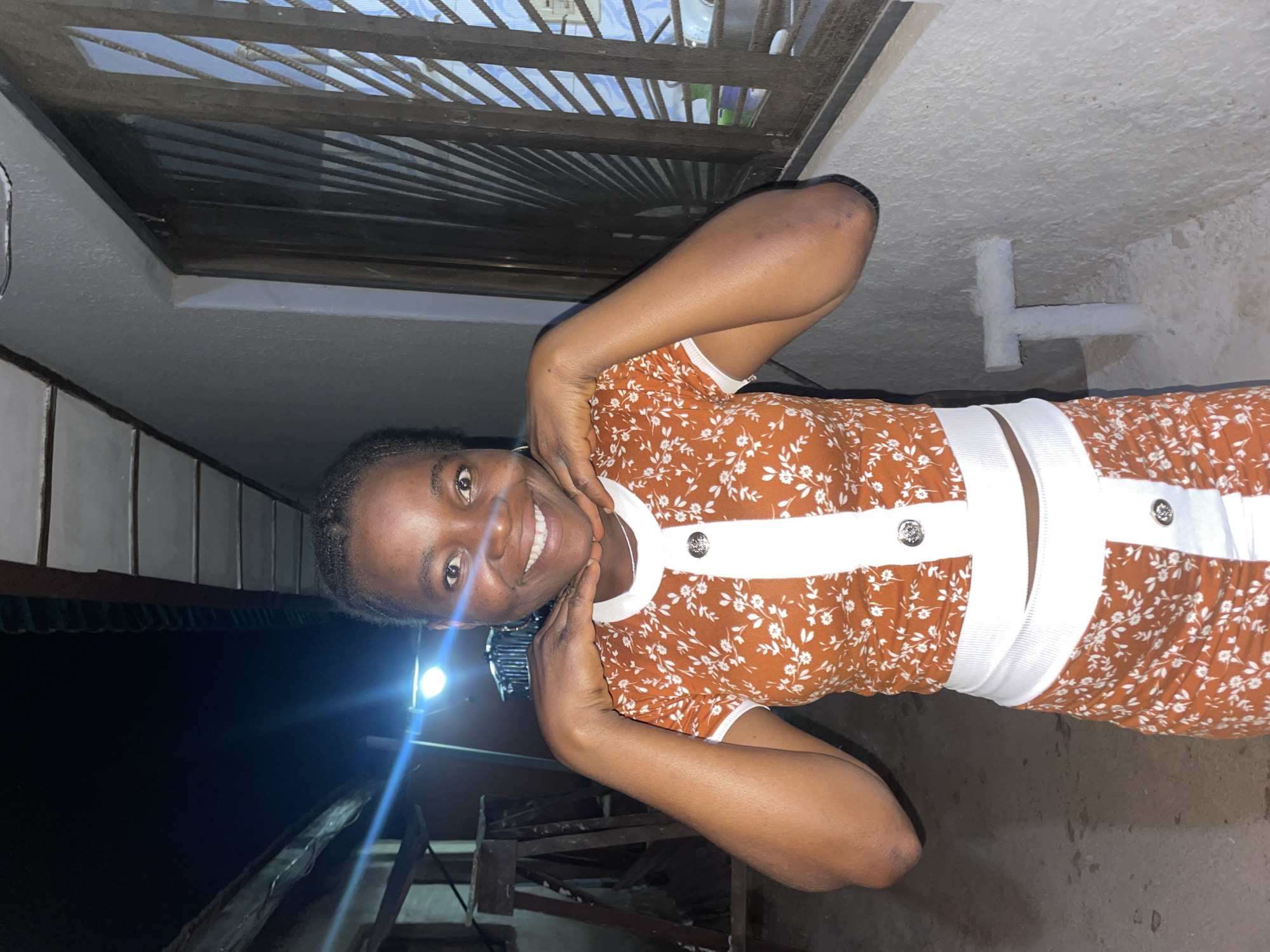 A photograph shows a young woman posing with her hands on her face while standing in a dimly lit location. The woman is wearing a floral print dress with white accents. Above her, a barred window is visible, and behind her, there's a dark, indistinct background with elements suggesting a building structure. A bright light source is positioned behind and to the left of the woman. The photo does not provide sufficient information to definitively determine the location in the city of Jos, Nigeria.