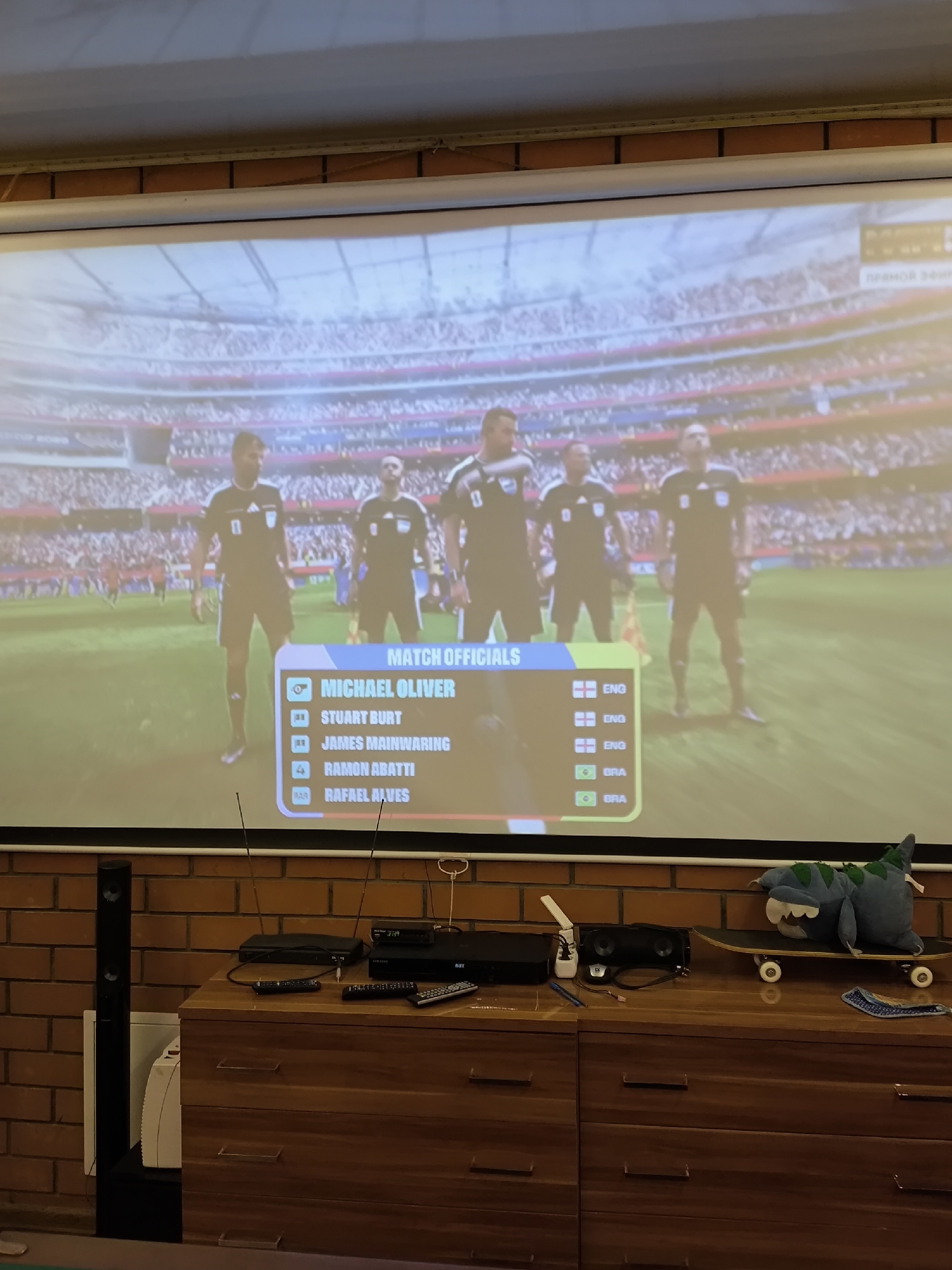 The image displays a projected screen showing a soccer video game. On the screen, there are four referees standing on a soccer field, with a list of "MATCH OFFICIALS" on the lower right. The list includes names such as Michael Oliver, Stuart Burt, James Mainwaring, Ramon Abatti, and Rafael Alves, along with small flag icons. The background shows a packed stadium. In the foreground, a wooden dresser is visible, with various electronic devices and remote controls placed on top. A black tower speaker is positioned to the left of the dresser. A plush toy dinosaur is resting on a skateboard on the dresser. The wall behind the dresser is made of brick. The overall setting suggests a home entertainment setup. The city of Pokrov, Russia is not depicted in the image.