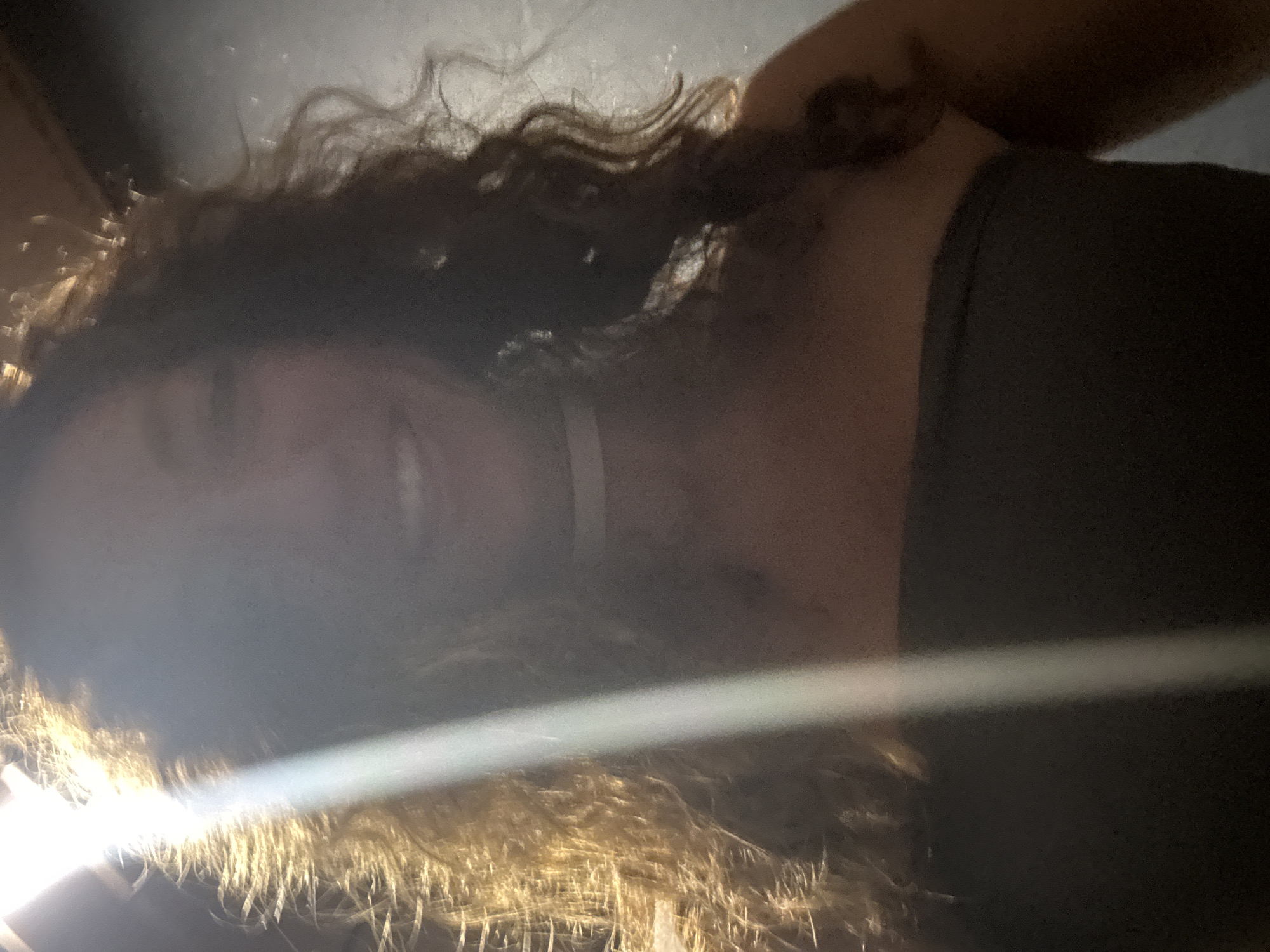 A person, likely female, is depicted in a portrait shot, presumably taken in Mù Cang Chải, Vietnam. A strong, bright light source from the upper left creates significant backlighting and lens flare, causing the left side of the subject's face and hair to be overexposed and appear golden, while the rest of their face and the background are cast in deep shadow. The subject has long, dark, curly hair and is smiling, revealing visible teeth. They are wearing a dark, strapless top and a white choker necklace. The background is uniformly dark and indistinct, lacking specific identifiable features.