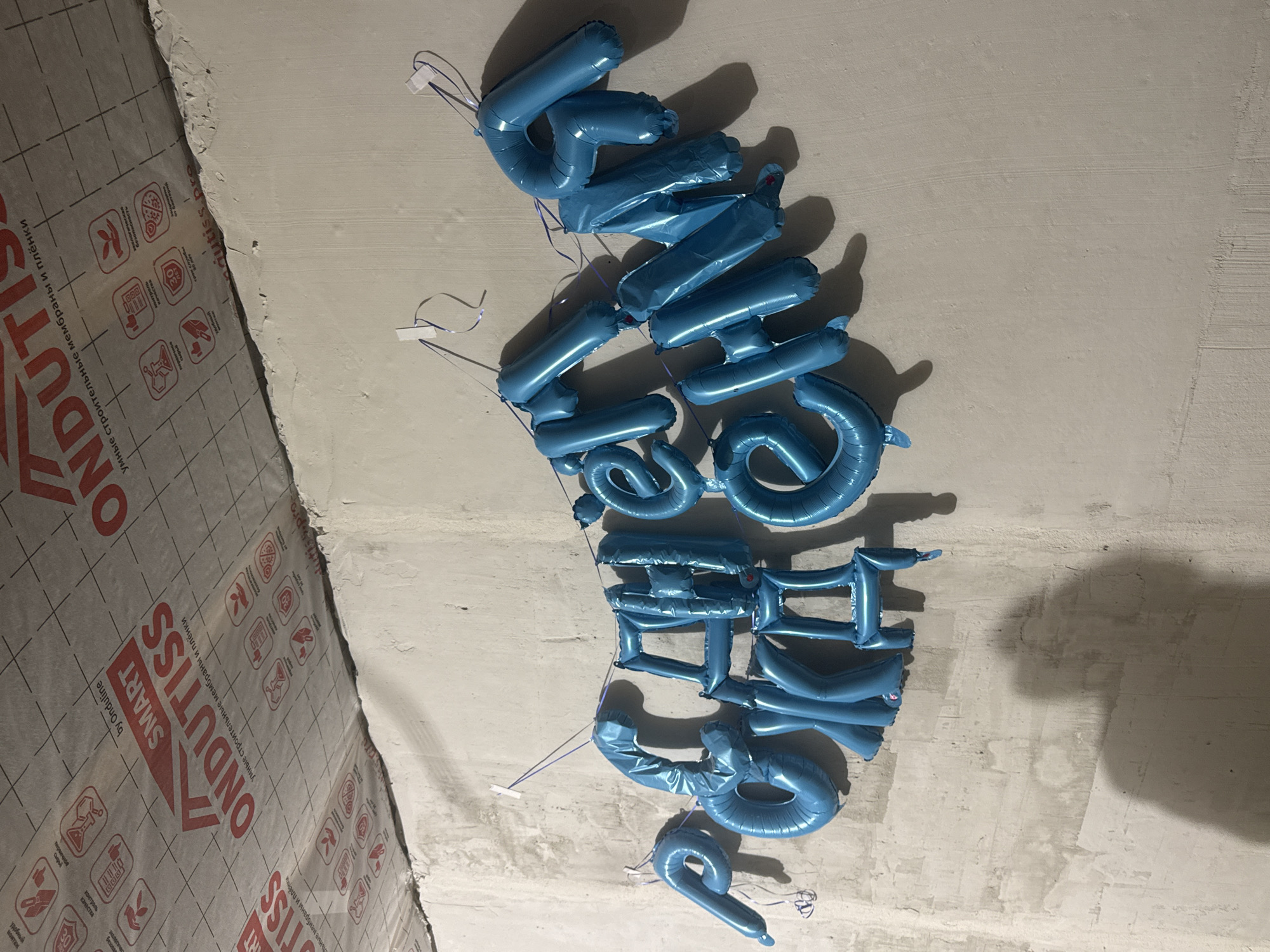 The image shows a wall with blue balloons forming the Russian text "С ДНЕМ РОЖДЕНИЯ", which translates to "Happy Birthday". The balloons are attached to the wall with purple ribbons. The wall appears to be in an unfinished building, as indicated by the concrete surface and exposed construction materials on the ceiling. No people are visible in the image. The context of Perm, Russia, is not discernible from the visual elements of the image itself.