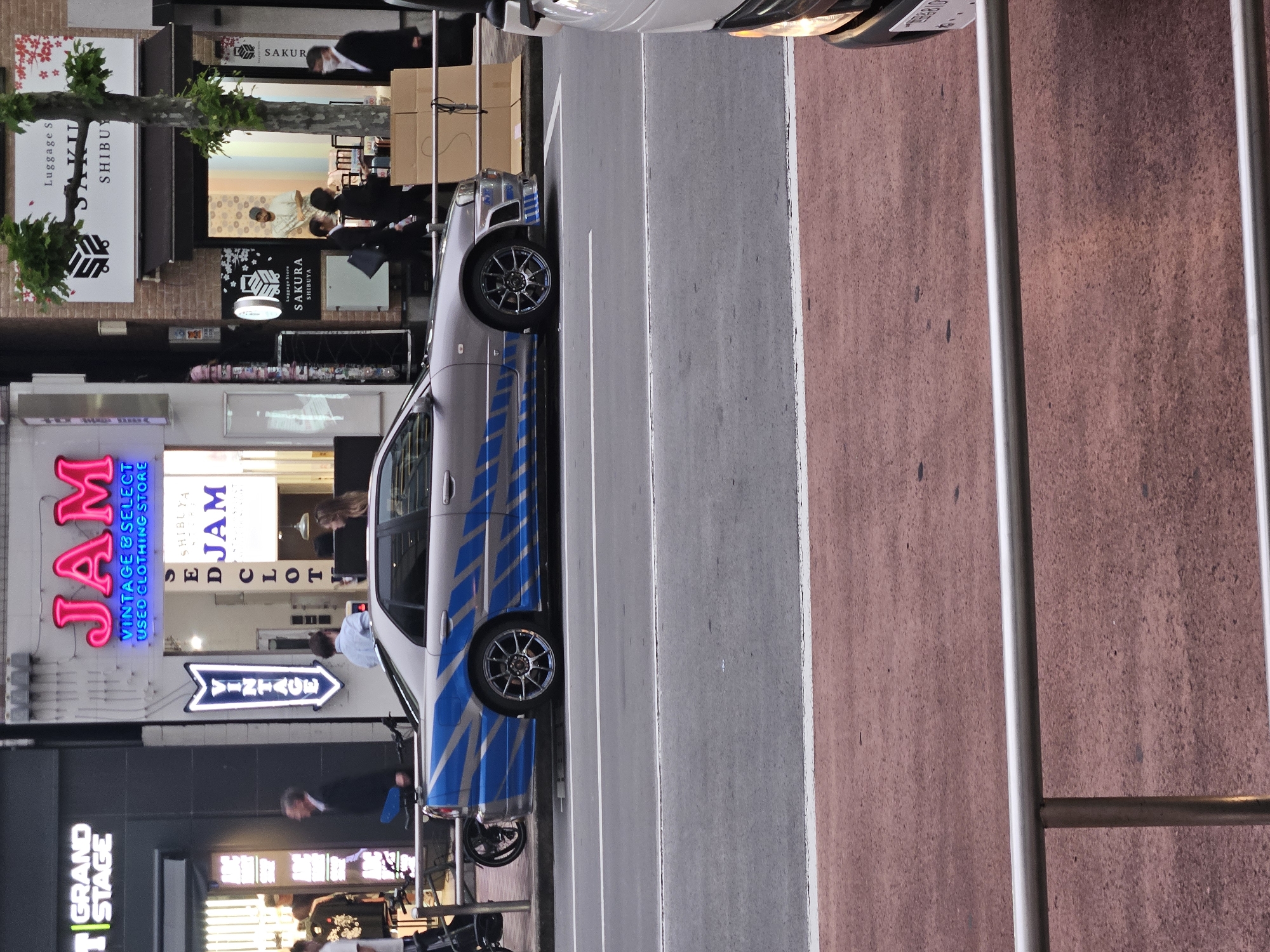 The scene depicts a street in Shibuya, Japan. Notable establishments visible are "Grand Stage," "JAM Vintage & Select Used Clothing Store," and "Sakura Luggage Store Shibuya." People are present, some appearing to be pedestrians. A silver car with blue stripes is parked along the street. The environment includes a street with pedestrian crosswalks. The buildings appear to be multi-story structures.