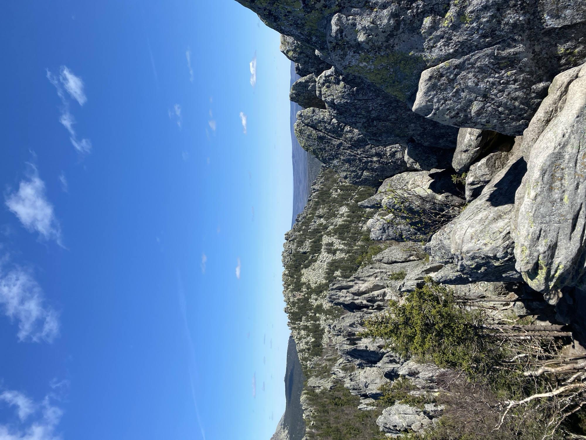 The image depicts a rocky landscape under a clear blue sky with scattered clouds. The terrain consists of gray rocks and sparse vegetation. The foreground shows lichen-covered boulders. In the distance, there is a view of tree-covered hills.
The location is in the city of Zlatoust, Russia, although without further information, the specific landmark or viewpoint is unidentifiable. There are no people visible in the scene.
