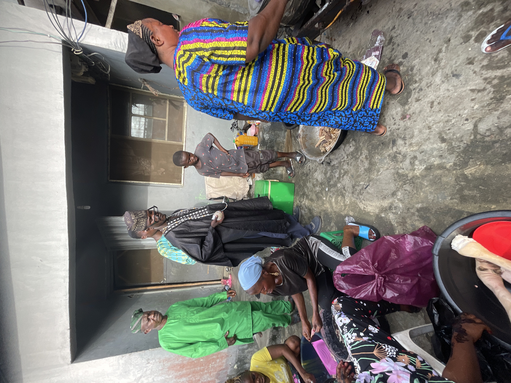 The scene depicts a group of people in Ado, Nigeria. One woman in a colorful blue, yellow, and pink striped dress stands near a metal bowl containing meat. A young boy wearing a patterned shirt and dark shorts stands nearby with his hand on his hip. Inside a building, a man in a black garment and kufi hat stands near a window with another man in a green outfit standing next to him. In the foreground, individuals are sitting and leaning over a large container containing what appears to be meat and other items. A purple plastic bag is visible nearby.
