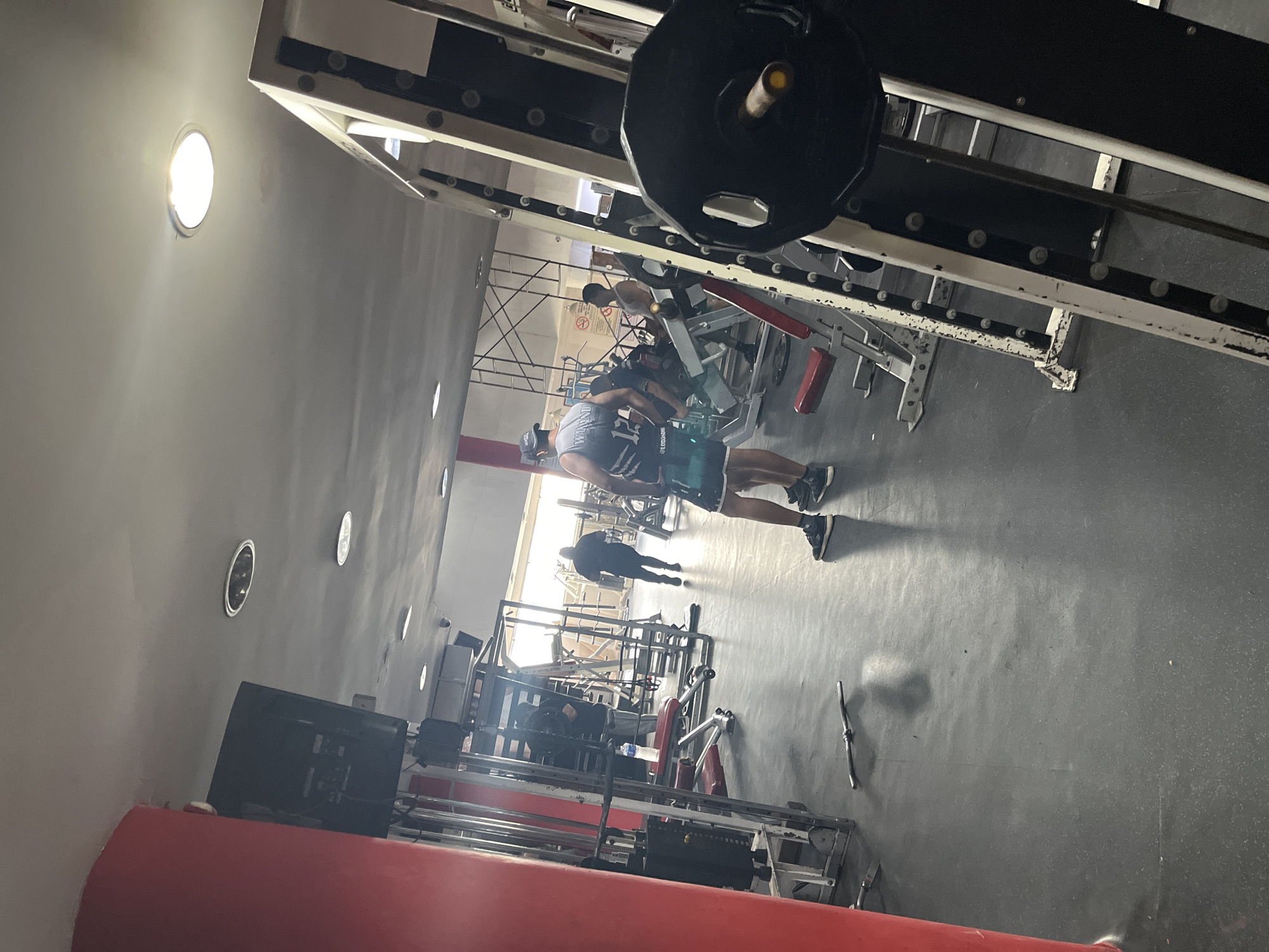 A medium, indoor shot in what appears to be a gym in Porlamar, Venezuela shows several individuals in the background. The gym has gym equipment such as weight machines. An individual is standing and talking to another individual with a mask on. There are lights that appear to be inset into the walls. A black weight is suspended in the top center of the image. The floor appears to be grey and smooth. The walls appear to be a flat grey, with a red support column. A window in the background shows bright sunlight.