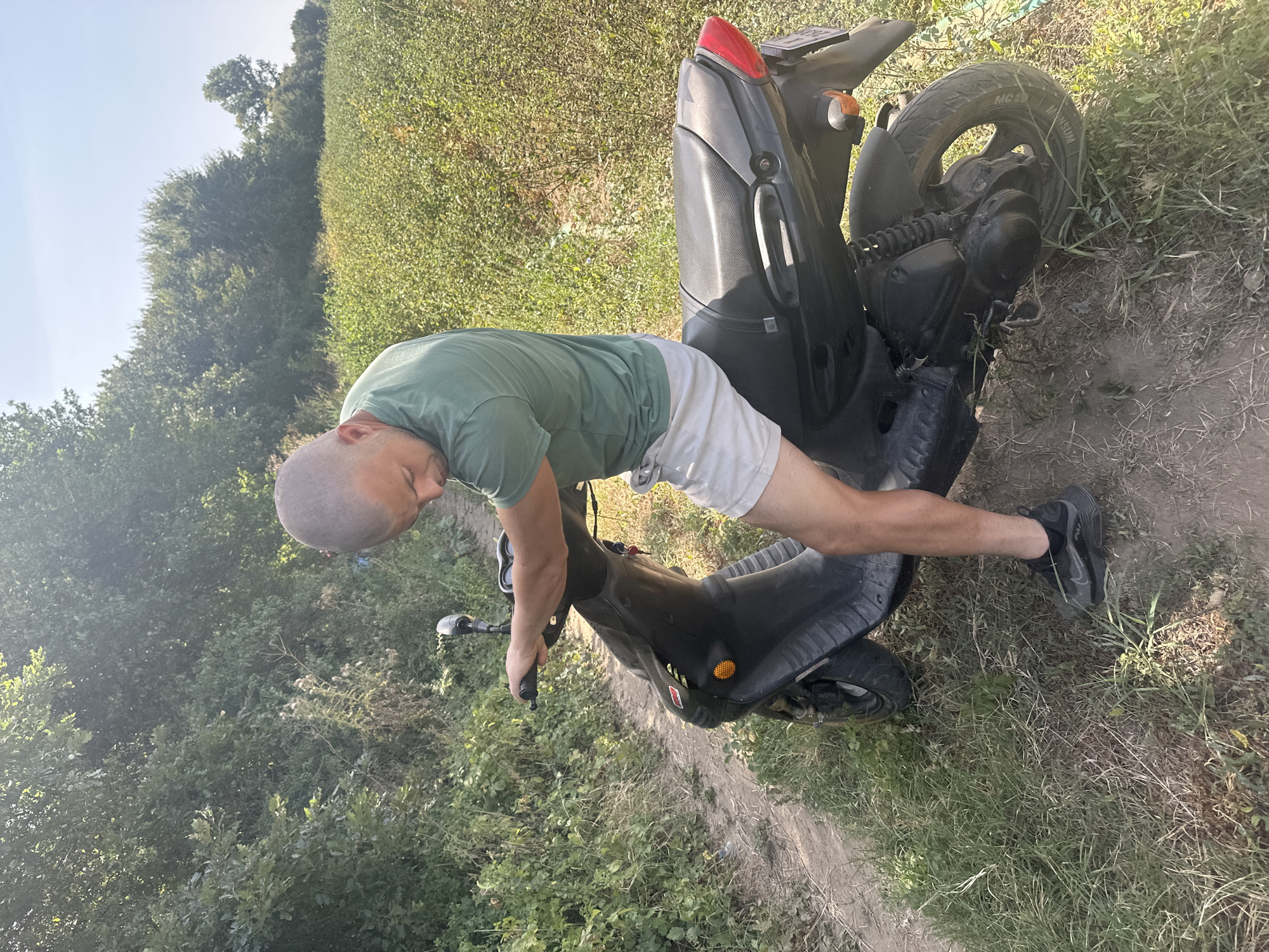 A man with a shaved head and a green t-shirt is sitting on a black scooter on a dirt path. The scooter is positioned at an angle, facing slightly to the left of the viewer. The man is leaning forward, with his right arm on the handlebars and his head down, looking towards the scooter. He is wearing light-colored shorts and black athletic shoes. The scooter has a red taillight and a license plate.
The environment appears to be rural. To the left of the path, there is dense green foliage and trees. To the right, there is a field of green plants under a bright, clear sky. The lighting suggests it is daytime, with sunlight casting shadows. The presence of the scooter and the dirt path implies an outdoor, possibly recreational or practical, setting. The location is identified as Malevo, Bulgaria.