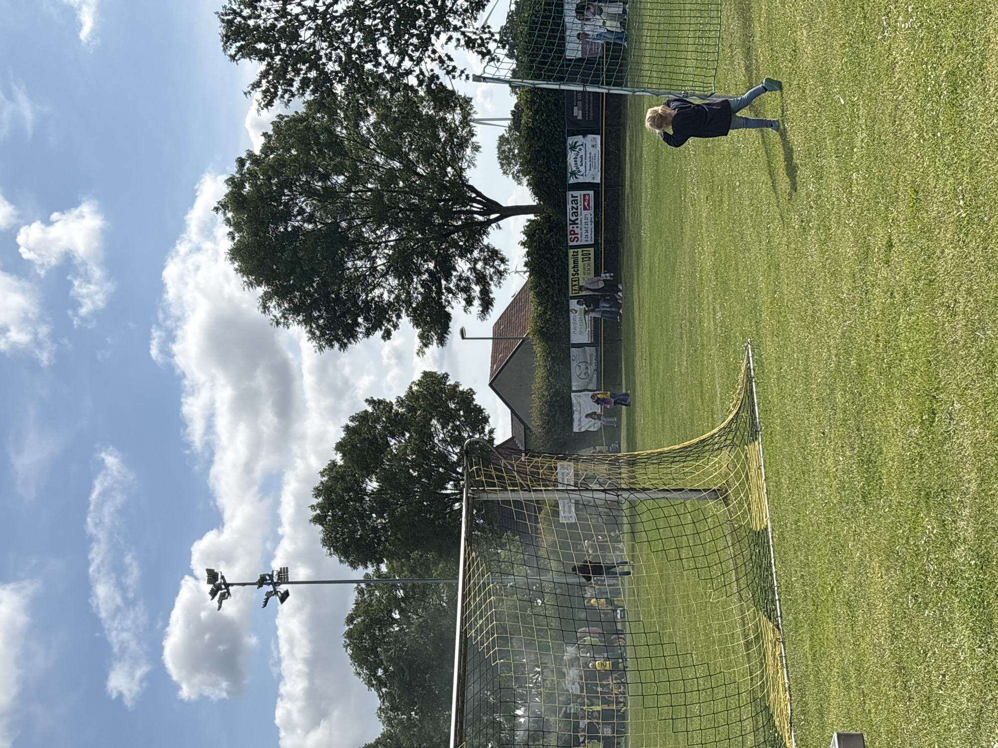 The image captures an outdoor scene at a soccer field in Wegberg, Germany, under a bright blue sky with scattered white cumulus clouds. In the foreground, a yellow and black soccer goal net with a white bottom frame is partially visible on the left. The field is covered in green grass. To the right, a young child with blonde hair, wearing a dark top, light blue jeans, and teal shoes, stands facing away from the viewer, looking towards the right, with their left arm slightly raised. The child casts a shadow on the grass.
In the midground, the expanse of the grass soccer field is visible, with several individuals and groups dispersed. On the left side of the field, beyond the foreground goal, a crowd of people, some wearing yellow shirts, appears to be gathered, partially obscured by a haze or dust. Further back, a long fence or barrier features multiple advertising banners. Legible text on these banners includes "TAXI SCHMITZ WEGBERG 0 24 34 / 1307", "SP:KAZAR 0 24 34 / 25 271 Mitte", and "Reisebüro Scholz". A group of people stands near the "TAXI SCHMITZ" banner. Another green net or goalpost is partially visible on the far right, with a few people standing behind it.
The background features dense green trees that line the perimeter of the field and partially obscure residential buildings with reddish-brown roofs. A tall floodlight pole with multiple lamps is prominently visible on the left side, extending into the sky. A faint, thin airplane contrail is discernible in the upper center sky.