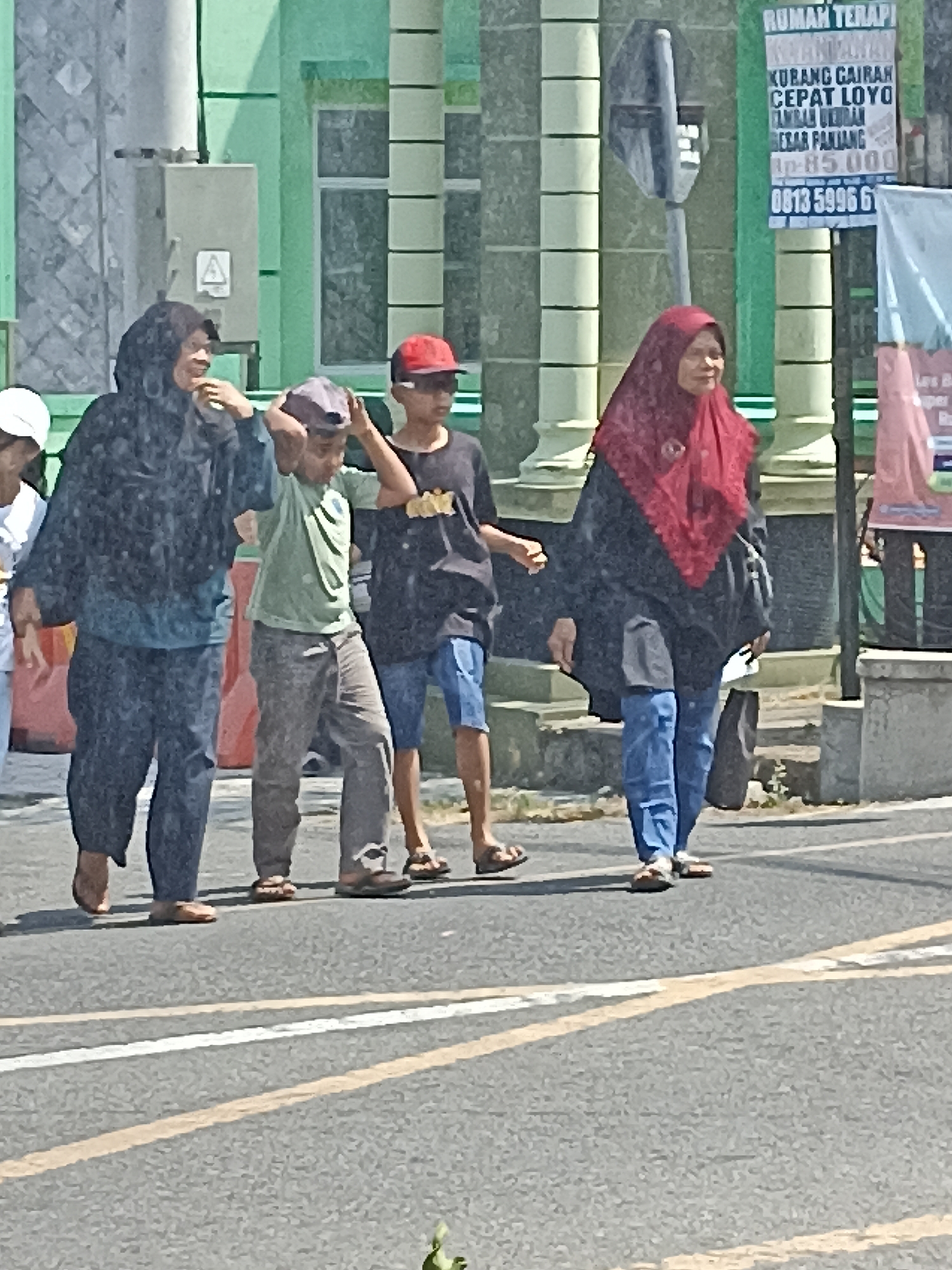 In Pulisen, Indonesia, a group of five people are walking on a street in front of a building with green walls and columns. The group consists of two women and three children. The woman on the far left is wearing a dark hijab and dark clothing. Next to her, a boy in a light green shirt and gray pants is adjusting his cap. A second boy, wearing a red cap and a black t-shirt with yellow lettering, walks beside him. The woman on the far right is wearing a red hijab and a dark top over blue jeans. A fifth person, mostly obscured, walks to the left of the first woman. The street has white and yellow lines. A sign with Indonesian text and numbers is visible on the building's exterior.