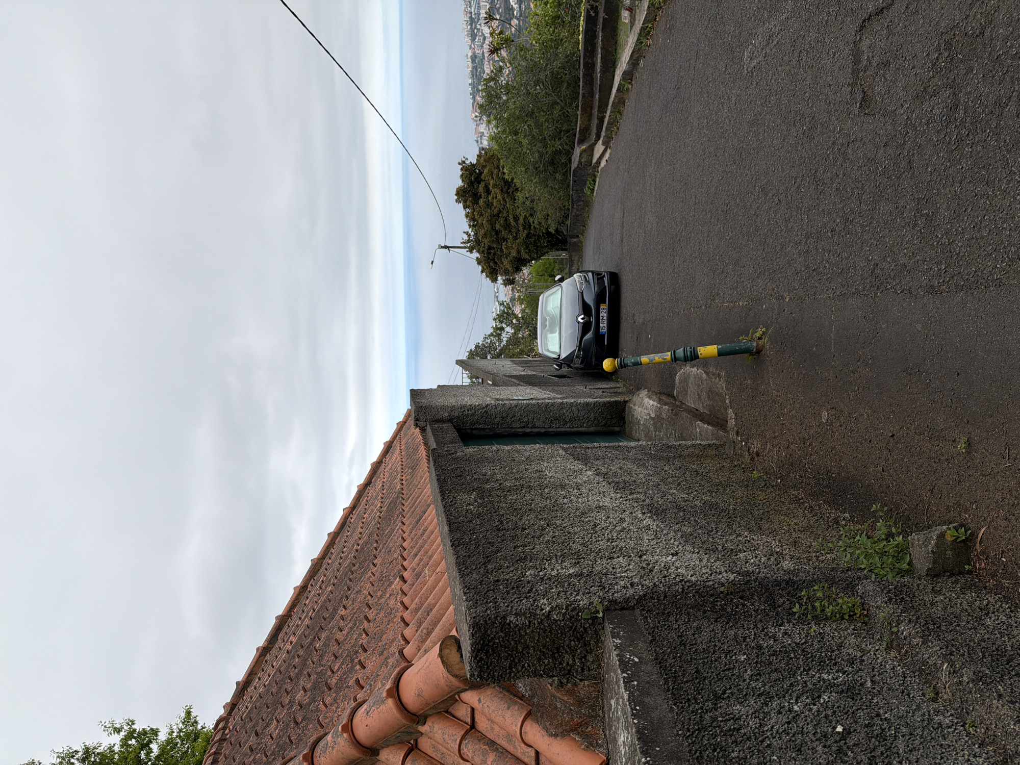A black car is parked on the side of a narrow paved road in Funchal, Portugal. The road slopes downward into the distance, with a view of the city and ocean visible in the background. A low stone wall lines the side of the road, with a section of terracotta roof tiles visible in the foreground. A green and yellow bollard stands beside the car. The sky is overcast.