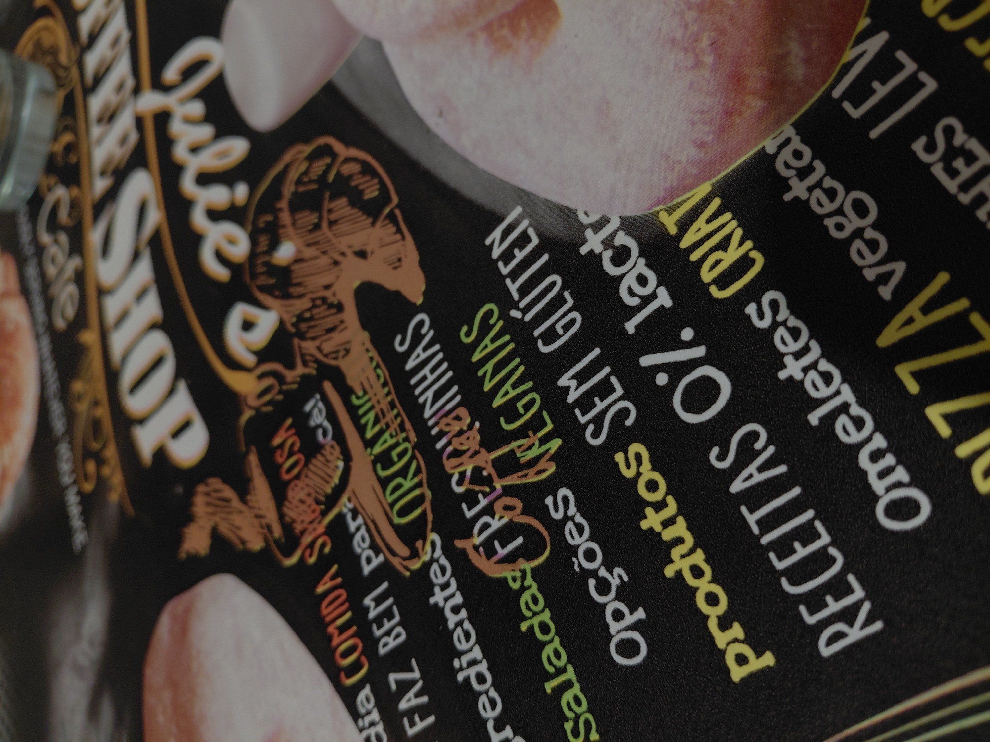 This detailed close-up image, captured in the city of Plovdiv, Bulgaria, presents a section of a black placemat or menu from "Julie's Coffee Shop." The text, primarily in Portuguese, is rendered in white, yellow, orange, and green fonts. Key phrases and categories visible include "Julie's Coffee Shop" in white script and block letters, followed by a list of offerings. These include "dia COMIDA SABOROSA para você!" (delicious food for you!), "FAZ BEM para você!" (it's good for you!), "Ingredientes ORGÂNICOS" (organic ingredients), "Saladas FRESQUINHAS" (fresh salads), "Opções VEGANAS" (vegan options), "Produtos SEM GLÚTEN" (gluten-free products), "RECEITAS 0% lactose" (0% lactose recipes), "Omeletes CRIATIVOS" (creative omelets), "PIZZA vegetariana" (vegetarian pizza), and "Lanches LEVES" (light snacks). Decorative orange illustrations, possibly depicting a figure or emblem, are integrated with the text. Blurred elements around the menu include a light-colored, possibly fruit, item (resembling a peach or nectarine) on the right, a small glass or container at the top right, and another light-colored, possibly baked good, object on the left. The scene implies an indoor cafe or restaurant setting.