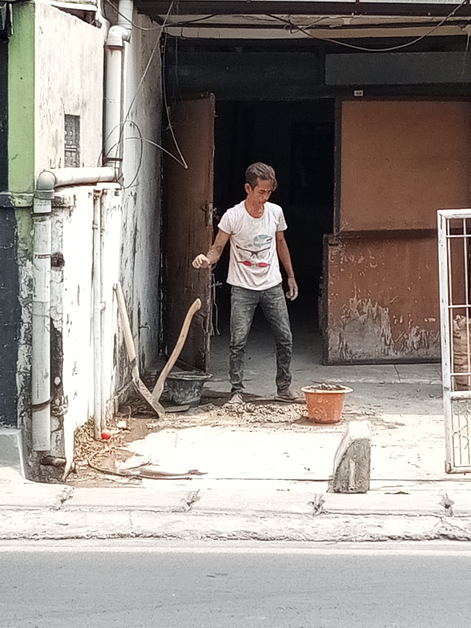A young man, appearing to be of Asian descent, stands in front of a building with an open doorway. He is wearing a white t-shirt with a graphic design and dark blue jeans. His right arm is raised, and his hand is extended as if reaching for something or interacting with the environment. He is positioned in what appears to be a construction or work area, with tools like a shovel and a bucket visible nearby. The ground is rough and uneven, with debris scattered around. The building itself shows signs of wear and tear, with peeling paint and exposed pipes on the exterior wall. The overall scene suggests a moment captured during a construction or renovation project in an urban setting in Cimahi, Indonesia.