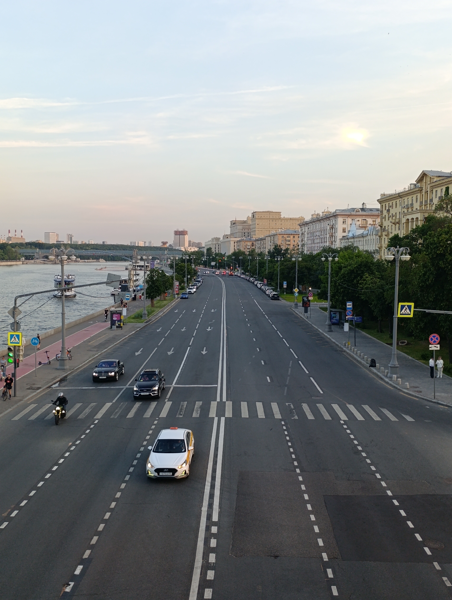 The image provides an elevated, wide-angle view of Tarasa Shevchenko Embankment in Moscow, Russia, looking generally west. The scene captures the late afternoon or early evening light under a partially cloudy sky.
On the left, the Moskva River flows calmly, featuring multiple large, multi-deck passenger boats docked along the red-paved cycle path and gray pedestrian walkway. Several people are visible strolling, cycling, or using scooters on the embankment. Trees line the riverbank. In the far distance on the left, industrial structures with tall cooling towers (likely a power plant, such as TEC-12) are visible, along with a bridge spanning the river.
The central part of the image is dominated by a wide asphalt road, Tarasa Shevchenko Embankment. It features multiple lanes delineated by white dashed lines, with a double solid white line separating traffic flows in the mid-ground. Lanes closer to the viewer show white directional arrows pointing away. Traffic includes a white Hyundai Solaris taxi (license plate "У671XM 45") and a motorcyclist approaching the viewer. Further away, a dark Bentley coupe and a dark Infiniti SUV are moving away from the camera. Numerous other cars are parked along the right side of the road in the distance. White-striped pedestrian crossings are marked across the road in the foreground and further along.
On the right side of the road, a wide sidewalk is lined with trees and streetlights. Various urban elements include a blue bus stop sign, a blue parking sign, yellow pedestrian crossing signs, and a red-on-blue "No Stopping" sign. The background on the right consists of a continuous row of multi-story buildings, characteristic of Stalinist-era architecture in Moscow, featuring light-colored facades and numerous windows. In the far distance on the right, a tall, modern building under construction, with a reddish-brown upper section, is visible, suggesting the proximity of the Moscow International Business Center (Moscow City).
Several pedestrians are present on the sidewalks and at crossings. Two individuals stand near the "No Stopping" sign on the far right. The sun or a bright light source creates a soft glow in the upper right portion of the sky.