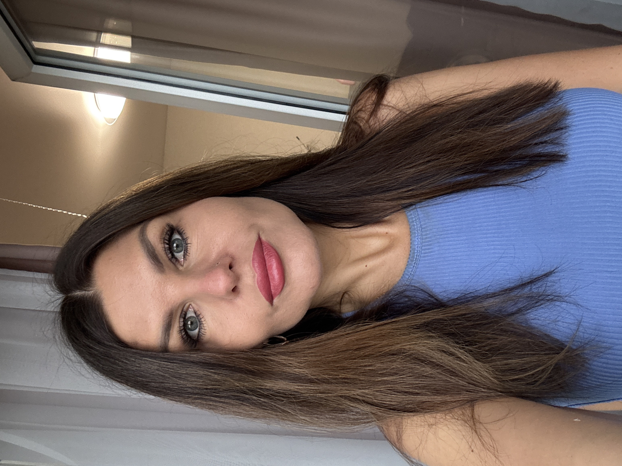 A woman with long brown hair is facing the camera. She is wearing a light blue top. She has blue eyes, pink lipstick, and a nose piercing. A window is visible in the background, with a light fixture above it. It is not possible to determine the location is Chaikovsky, Russia from the image.