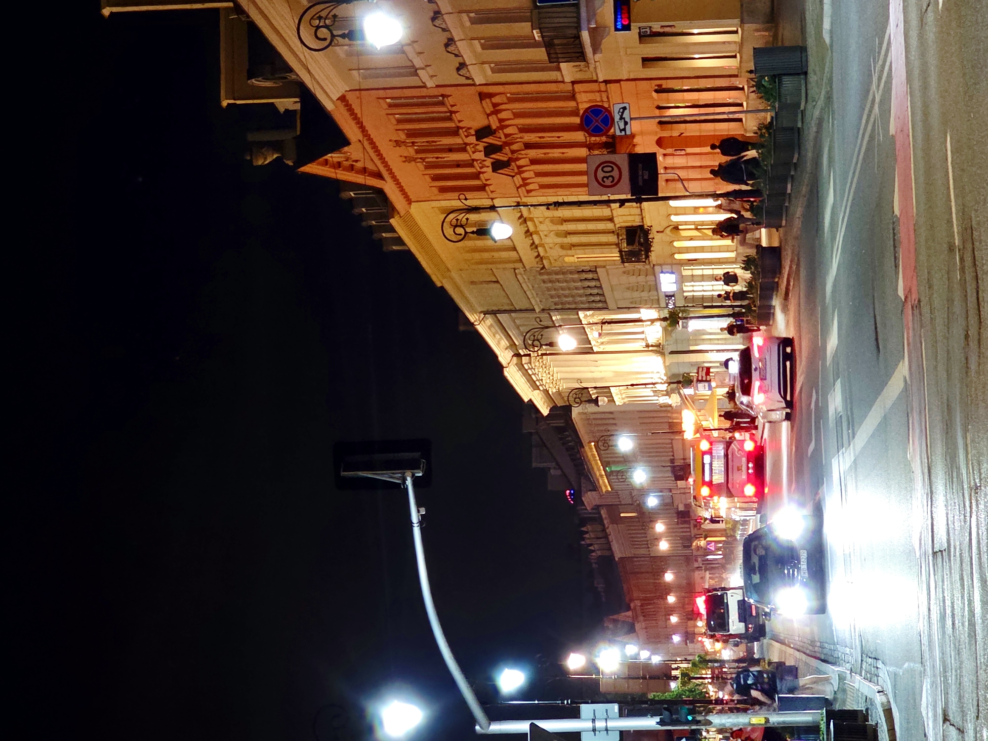 The image captures a nocturnal street scene in Warsaw, Poland, likely along a busy boulevard with illuminated buildings and traffic. On the left, a traffic light pole with a curved arm stretches across the street, illuminating the scene with a bright light. Further down the street, a white truck with its lights on is visible, followed by a red and white bus. In the foreground, a dark-colored car with its headlights on drives down the wet street, its lights reflecting on the road. Several pedestrians are seen walking along the sidewalks, with some gathered near planters. The buildings lining the street are a mix of styles, some appearing to be old architecture with ornate details, while others are more modern, all lit up and casting a warm glow. Signage indicating traffic regulations, such as a "30" speed limit sign, are present. The overall atmosphere is that of an active urban environment at night, with streetlights, vehicle lights, and building lights creating a vibrant and somewhat reflective scene on the wet pavement.