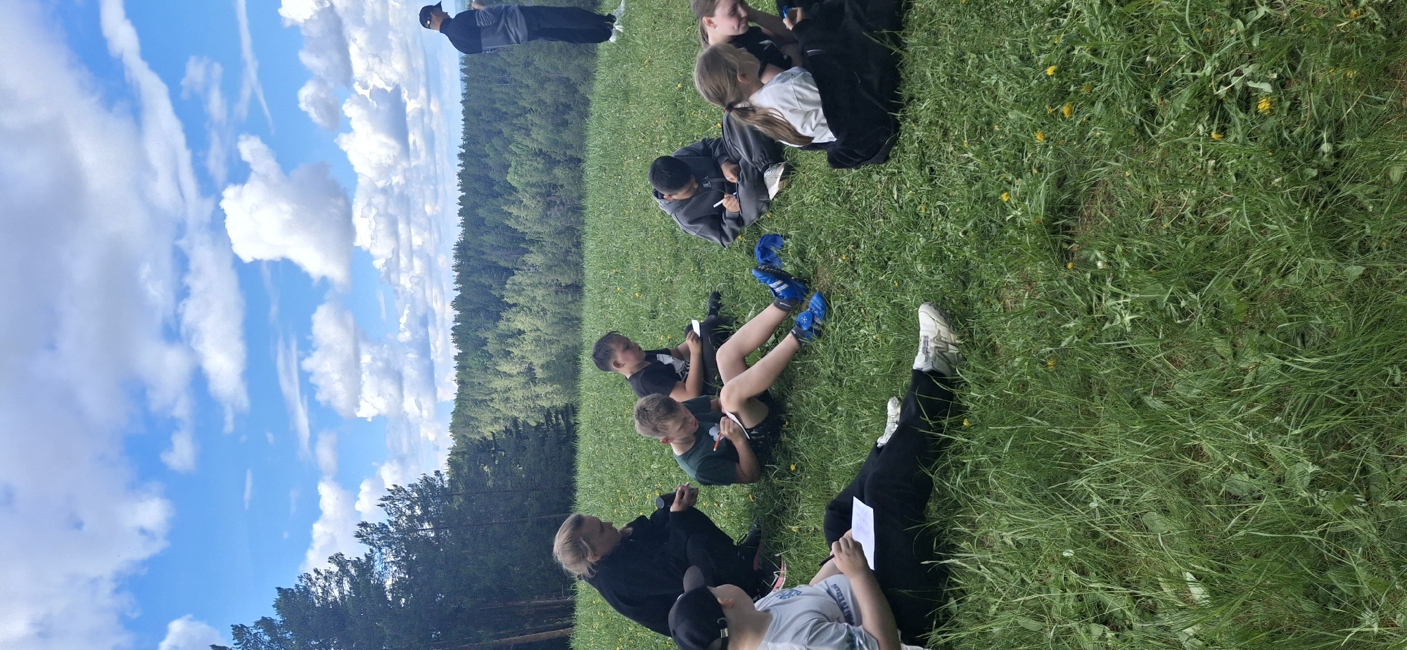 The image depicts a group of people, mostly children, sitting on a grassy field. They appear to be engaged in a writing or drawing activity, as many of them are holding notebooks or papers and writing utensils. The background includes a dense forest and a blue sky with scattered clouds. An adult in dark clothing is standing in the background overseeing the children. Based on the location information provided, this scene could be taking place in or near Прикамский, Russia.