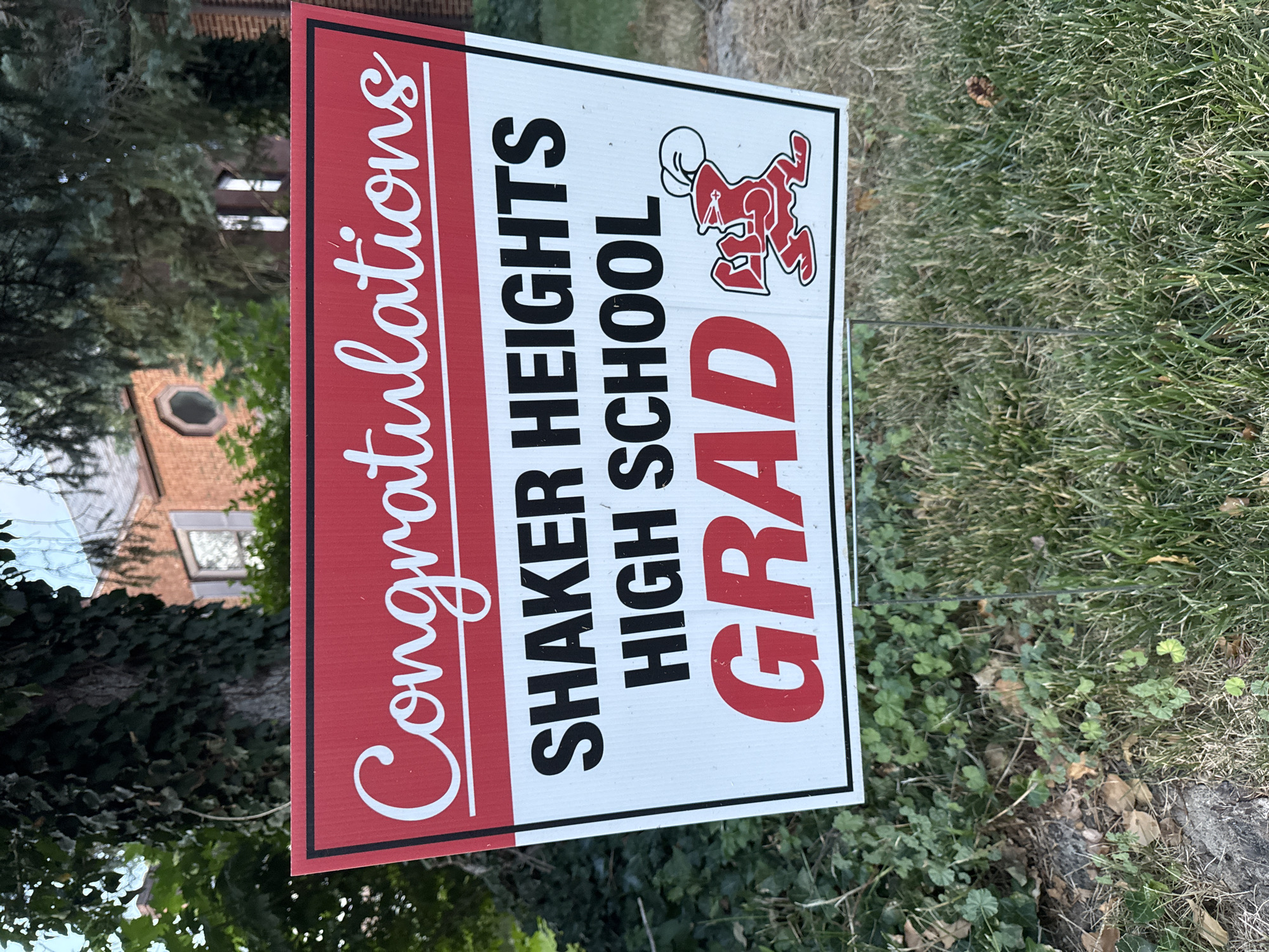 A celebratory yard sign for a graduate of Shaker Heights High School is displayed on a lawn. The sign features the text "Congratulations" in cursive white lettering on a red banner at the top. Below this, in bold black lettering, are the words "SHAKER HEIGHTS HIGH SCHOOL." The word "GRAD" is prominently displayed in large red capital letters at the bottom, accompanied by a red cartoon mascot with a chef's hat. The sign is supported by metal stakes and is situated in front of a brick house with a pitched roof and a visible circular window. Lush green trees and ivy are visible in the background, along with a patch of grass and some fallen leaves in the foreground. The scene appears to be outdoors in a residential setting.