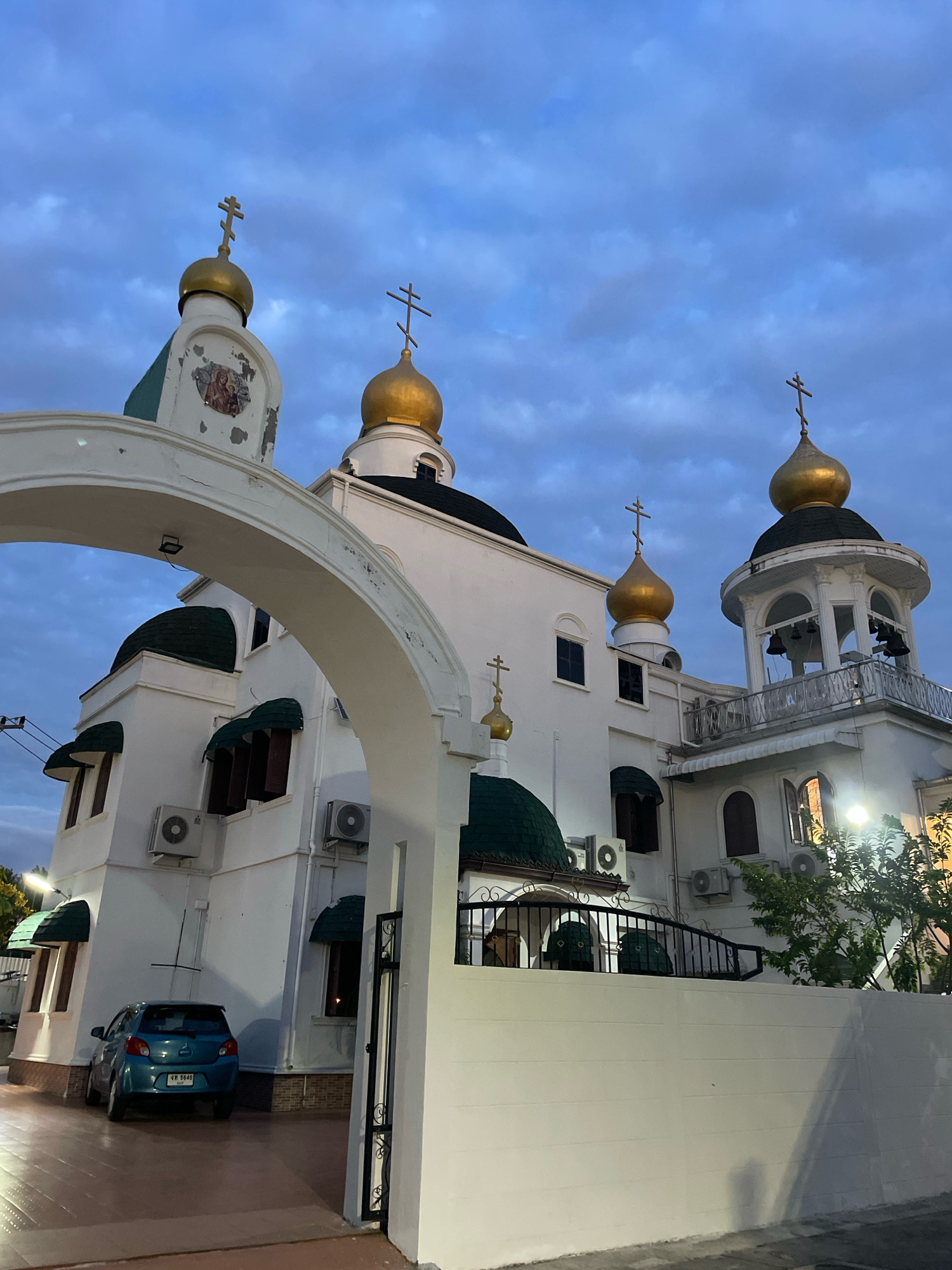 The image captures an exterior view of the Orthodox Christian Church of All Saints in Pattaya City, Thailand, at dusk. The sky is a deep blue with scattered clouds. The church is predominantly white with multiple golden domes topped with crosses. Some domes are green. An arched entryway is visible in the foreground, framing the view of the church. Air conditioning units are mounted on the exterior walls of the church. A small blue car is parked on a paved driveway to the left. A black metal fence runs along the front of the property. A bell tower with bells is visible on the right side of the church structure. Trees and foliage are present to the right of the church. The overall scene is serene and architectural.