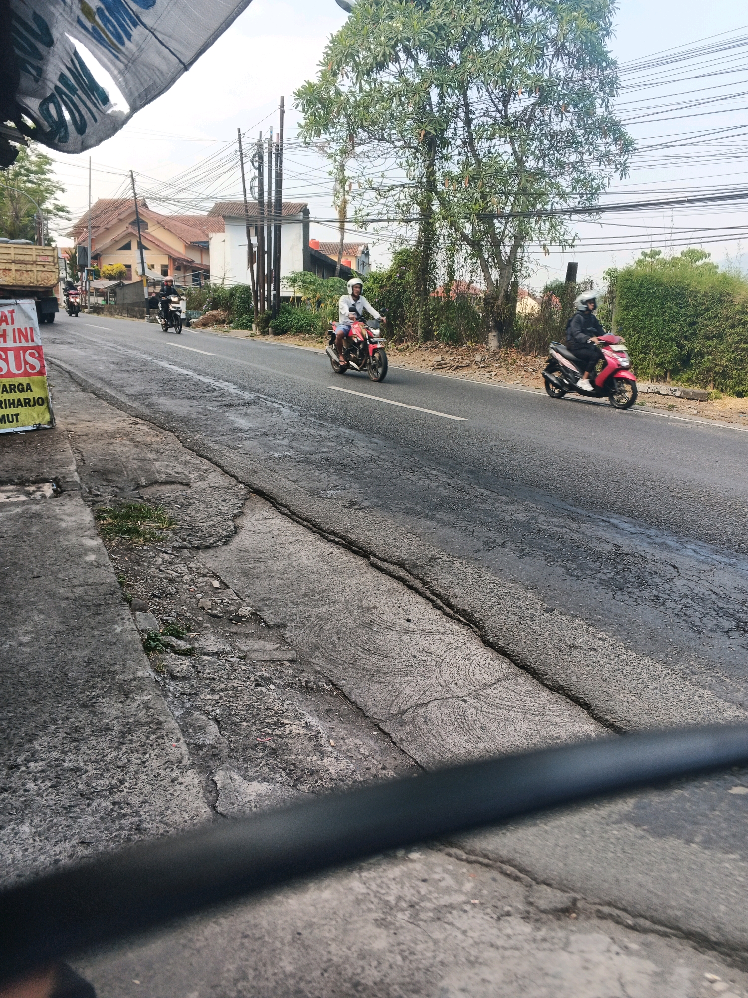 A street scene in Ungaran, Indonesia, features several motorcycles traveling on a paved road. In the foreground, a damaged concrete sidewalk is visible, with cracks and uneven surfaces. A tall, leafy tree stands on the right side of the road, and behind it, dense green foliage lines the roadside. In the background, houses with reddish-brown tiled roofs are nestled amongst more trees. Overhead, a complex network of electrical wires crisscrosses the sky. A large, yellow truck with a cargo bed is parked on the left side of the road, with a sign on its side partially obscured by a white, tattered banner. The sky is overcast, suggesting either early morning or late afternoon. One person on a red motorcycle wears a white helmet and blue shorts, while another on a pink scooter is dressed in black with a white helmet.