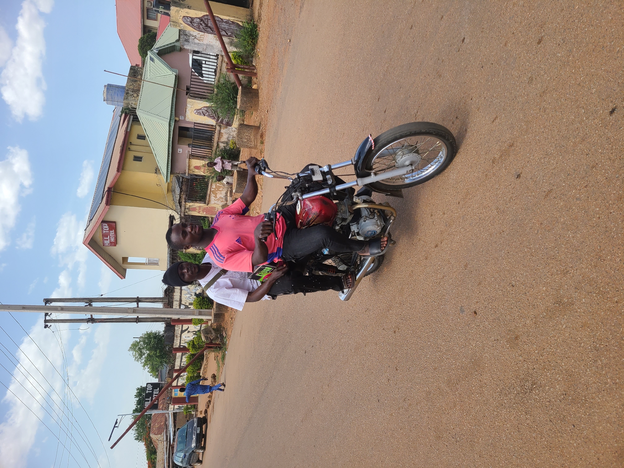 Two people are riding a motorcycle on a paved road. The rider is wearing a pink shirt and the passenger a white shirt and black hat. The buildings nearby are two-story with a sign reading "Hill Top Hotel". The location is a city in an Unknown region.