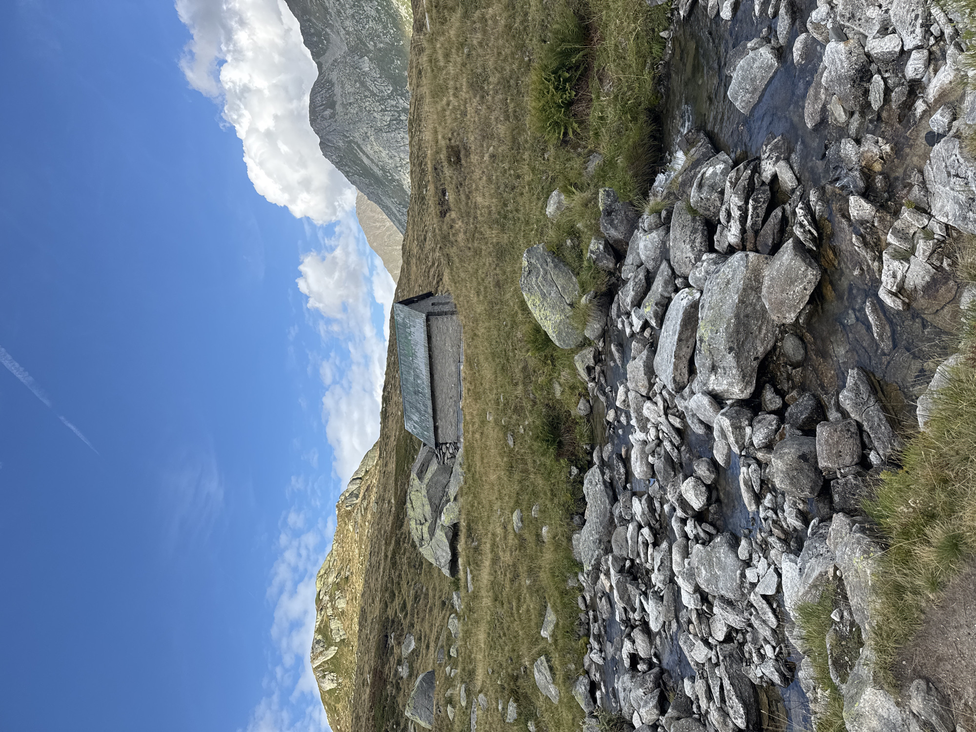 The image depicts a serene mountain landscape, likely in the vicinity of Airolo, Switzerland, characterized by a rustic stone hut with a green roof situated on a grassy slope. A rocky stream flows in the foreground, its clear water weaving through large, grey stones and sparse vegetation. The background features rolling hills and majestic mountains under a partially cloudy blue sky, with a faint contrail visible. The scene is devoid of human presence, emphasizing the solitude and natural beauty of the alpine environment. The overall impression is one of tranquility and rugged, untouched wilderness.