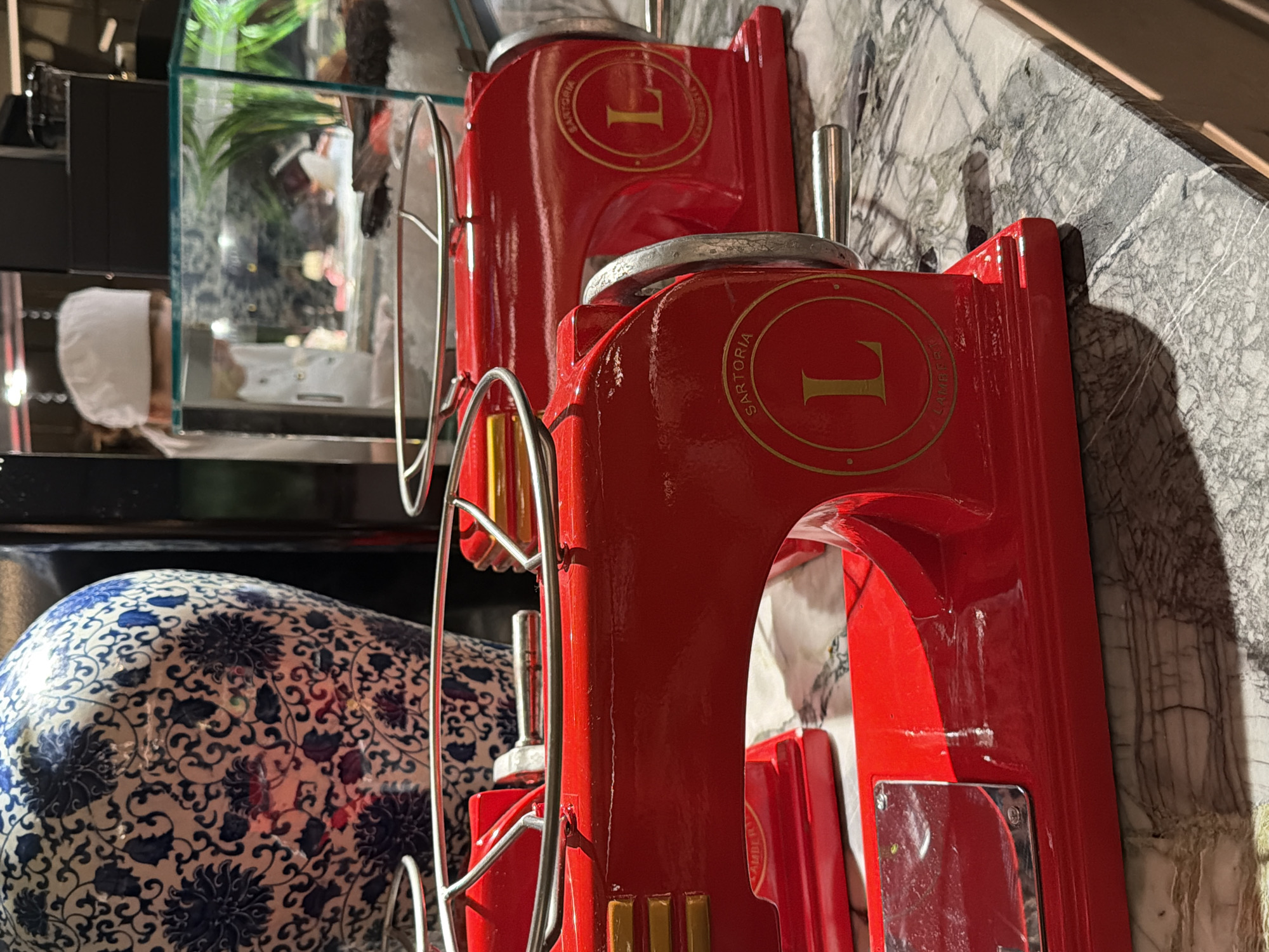 The image, oriented vertically, depicts an indoor scene likely within a high-end restaurant or food service area in Moscow, Russia. Dominating the foreground are two vibrant, glossy red machines, possibly industrial-style food slicers or display units, resting on a polished gray and white veined marble countertop. Each machine features metallic silver components and a golden circular emblem prominently displaying the number "1" in its center. The lower machine clearly shows the text "SARTORIA" inscribed around the golden circle.
In the background, a person wearing a white chef's hat or head covering is partially visible behind a glass partition or display case, suggesting they are working or preparing items. The glass case contains green plants and other indistinct objects, possibly food, contributing to a professional culinary environment. To the lower left, a portion of a large, decorative ceramic vase with an intricate blue and white floral pattern (chinoiserie style) is visible, adding an elegant touch to the setting. The scene is brightly lit, with reflections on the glossy surfaces of the red machines and the marble countertop. No specific activity is clearly unfolding beyond the general operational ambiance of a food establishment.