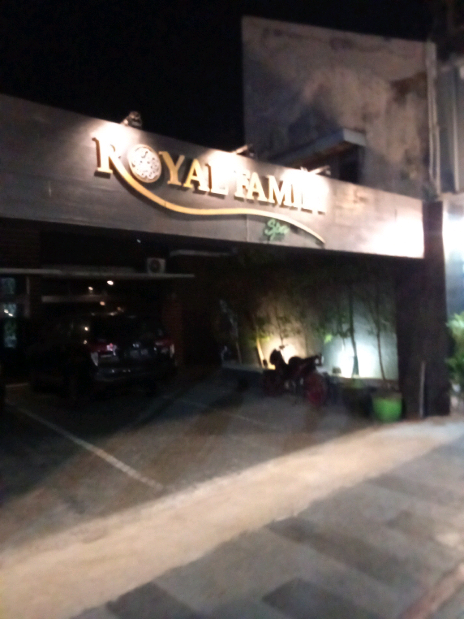 A dark, outdoor scene at night, likely in Kediri City, Indonesia, shows a building entrance with a prominent sign reading "ROYAL FAMILY" in gold lettering. Below the main sign, smaller green text appears to read "Spa". A black SUV is parked under the covered entrance. To the right of the entrance, a motorcycle is parked, and beyond it, there is a large green planter and the facade of a building. The ground is paved. The lighting suggests artificial illumination from the building and possibly streetlights.