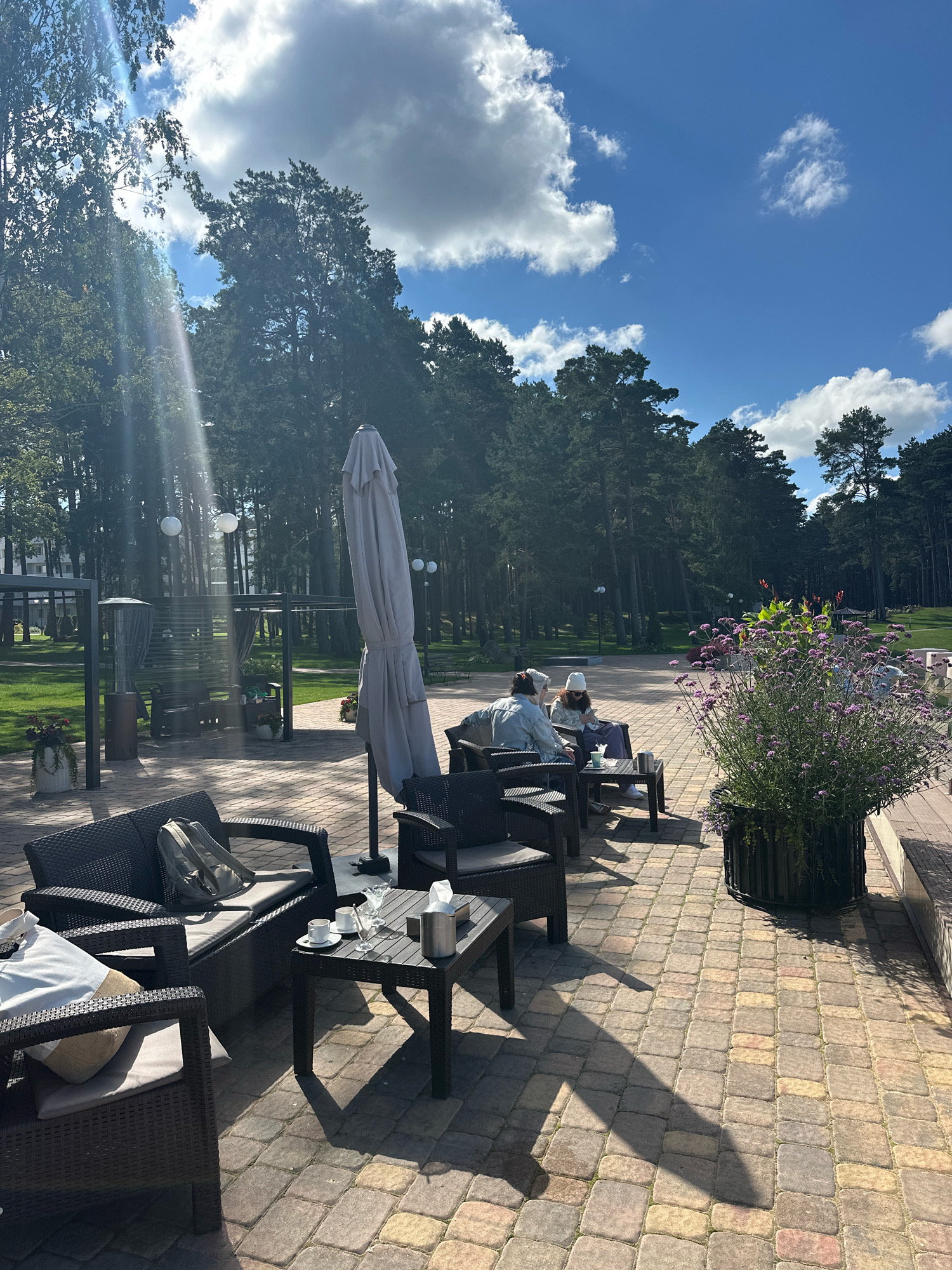 In an outdoor seating area of a establishment located in the Мядзельскі сельскі Савет, Belarus, two individuals are seated on dark wicker furniture. One person is wearing a white hat and light-colored clothing, while the other wears a light blue jacket and dark pants. They are positioned at a small black table where two cups of coffee and glasses are present. In the foreground, another dark wicker sofa holds a grey backpack and cushions.
The area is paved with multi-colored rectangular stones. A large, light-colored umbrella is folded and positioned vertically to the right of the seating area. Beyond the patio, a manicured green lawn is visible, with trees lining the background. Sunlight beams through the trees, creating lens flare and casting shadows on the patio. A large potted plant with purple flowers is situated to the right of the table. In the middle ground, a covered seating structure with hanging lights and a patio heater can be observed. The sky above is blue with scattered white clouds.