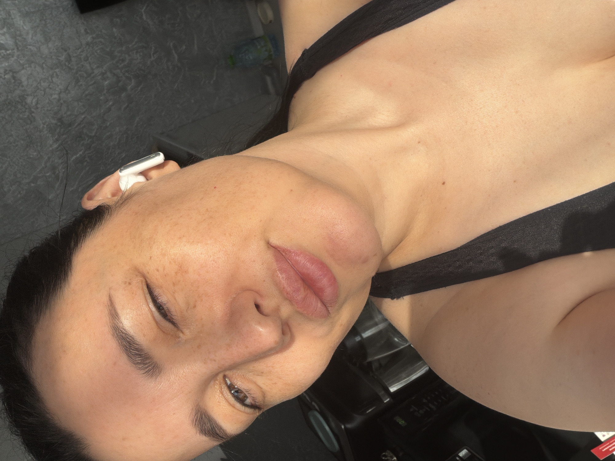 A close-up selfie shows a woman winking and puckering her lips, with a white wireless earbud in her right ear. She is wearing a black sports bra. The lighting suggests an indoor setting, possibly a home, with sunlight streaming in from the side, casting shadows and highlighting her freckles. The background is blurred, showing a dark, textured wall and some indistinct objects that could be kitchen appliances or furniture. The woman's skin appears to be lightly tanned and her hair is dark and pulled back. The overall impression is one of casualness and self-expression.