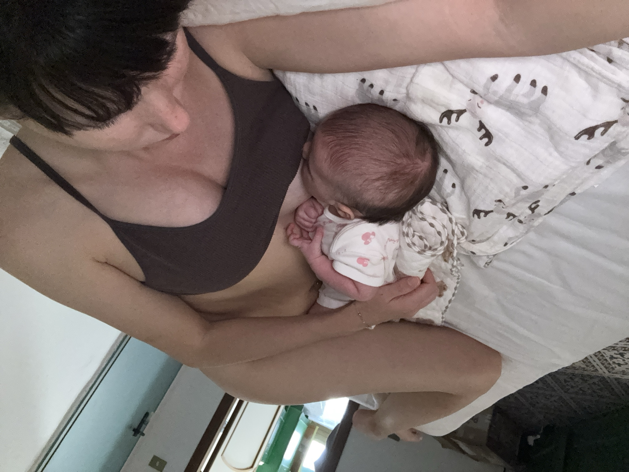 The image shows a close-up, overhead view of a person and a newborn baby lying on a bed. The person, whose face is partially visible and obscured by their dark hair, is wearing a dark brown bra and appears to be breastfeeding the baby. The baby, facing away from the camera and towards the person, is wearing a white onesie with pink designs. The baby's head is visible, as are their tiny hands. They are lying on a white, patterned swaddle blanket. The background suggests an indoor setting with natural light streaming in from a window. The overall scene is intimate and peaceful, focusing on the bond between the person and the infant. No specific landmarks or contextual clues place the scene within Isola delle Femmine, Italy, other than information provided separately.