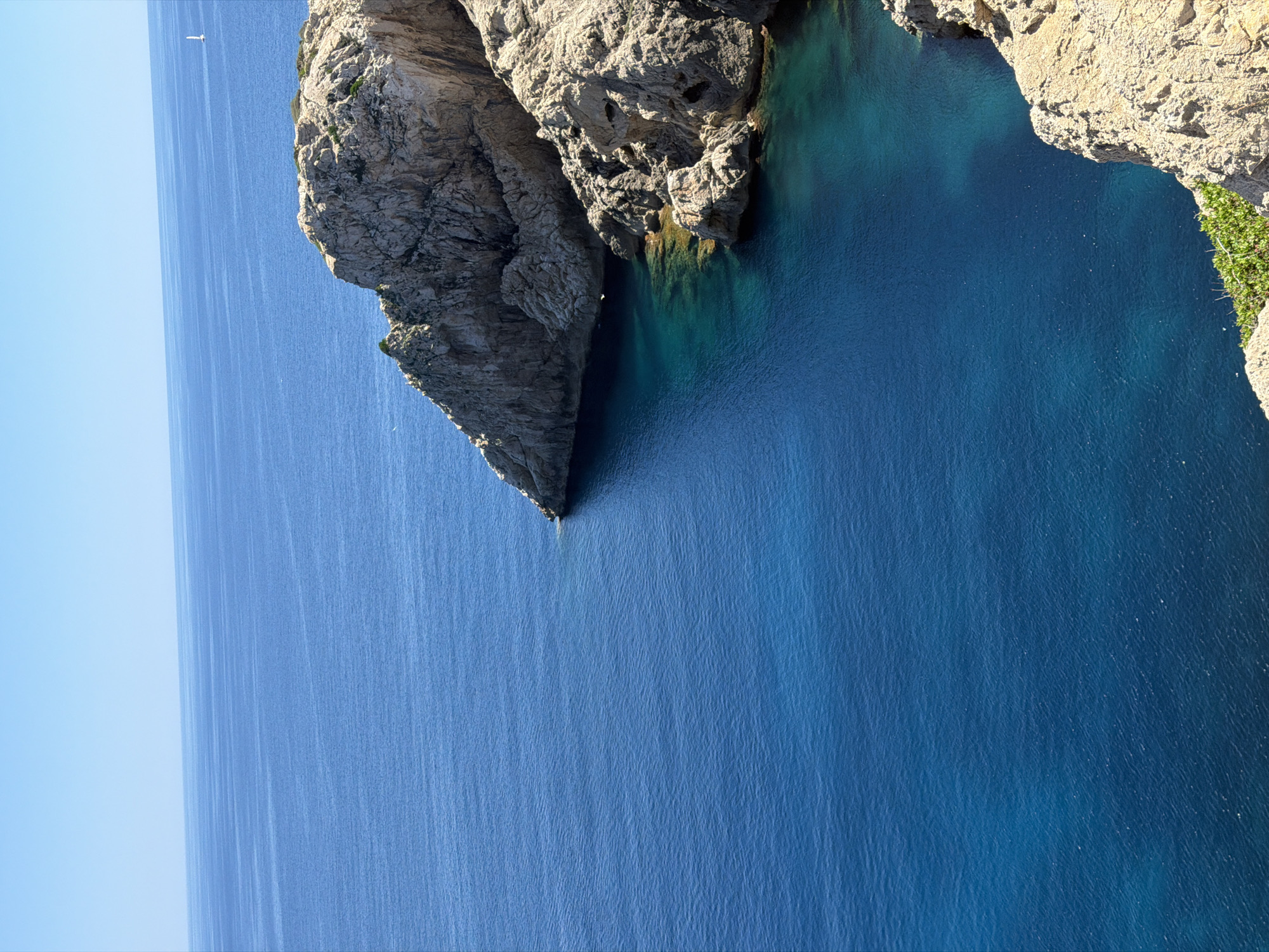 A coastal scene is presented with a rocky outcrop featuring a triangular spit of rock extending into the water. The ocean transitions from a lighter azure to a deep blue. At the top of the image, a light, rocky cliff edge is visible, with some green vegetation clinging to it. The location is Cala Gat, Spain.