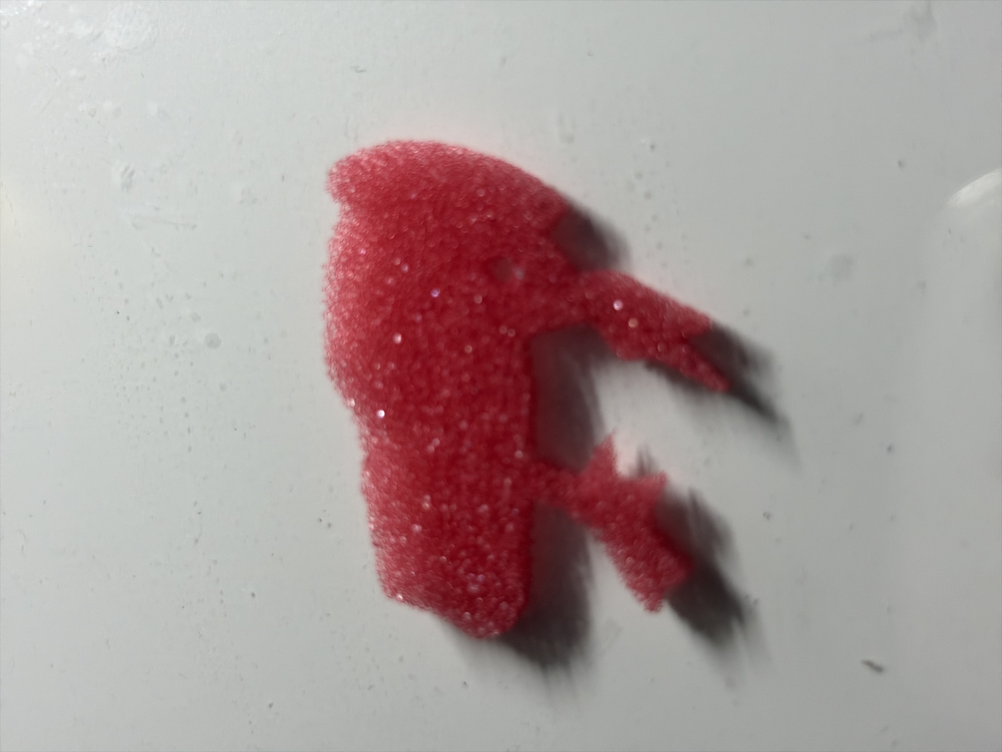 A small, bright pink, glittery object shaped like a stylized bison is positioned on a white, textured surface. The surface appears to be a tabletop or counter, with some faint water spots or smudges visible. The bison-shaped object is the sole focus of the image and is centrally located. There are no people, buildings, or any discernible Moscow landmarks present in the image.