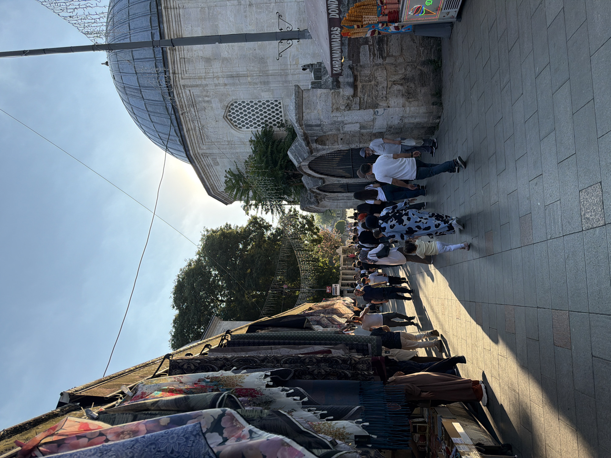 The image depicts a street scene in Eyüpsultan, Turkey, with a mosque visible on the right and market stalls lining the left. The sky is clear and blue with the sun casting long shadows across the paved walkway. A crowd of people, dressed in a variety of casual clothing, are walking in both directions. On the left, textiles are displayed for sale on stalls. The mosque has a prominent dome and stone facade. An ice cream cart is visible on the right side of the street.