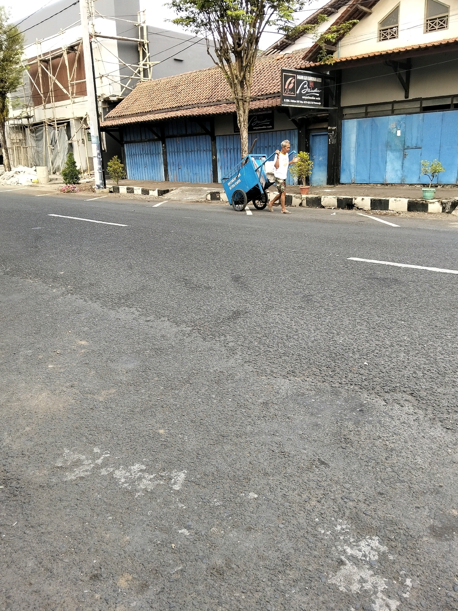 A man is pushing a blue cart along the side of a road in Purworejo, Indonesia. He is wearing a white t-shirt and shorts. The cart has two large wheels in the back and two smaller wheels in the front. The man appears to be walking from left to right in the image. The background shows a building with blue shutters and a sign that says "Digenggang Karu Banguunan & Bess." There are also trees and potted plants visible. The road is paved with asphalt and there are white lines painted on it. The scene is set during the daytime, with good lighting.