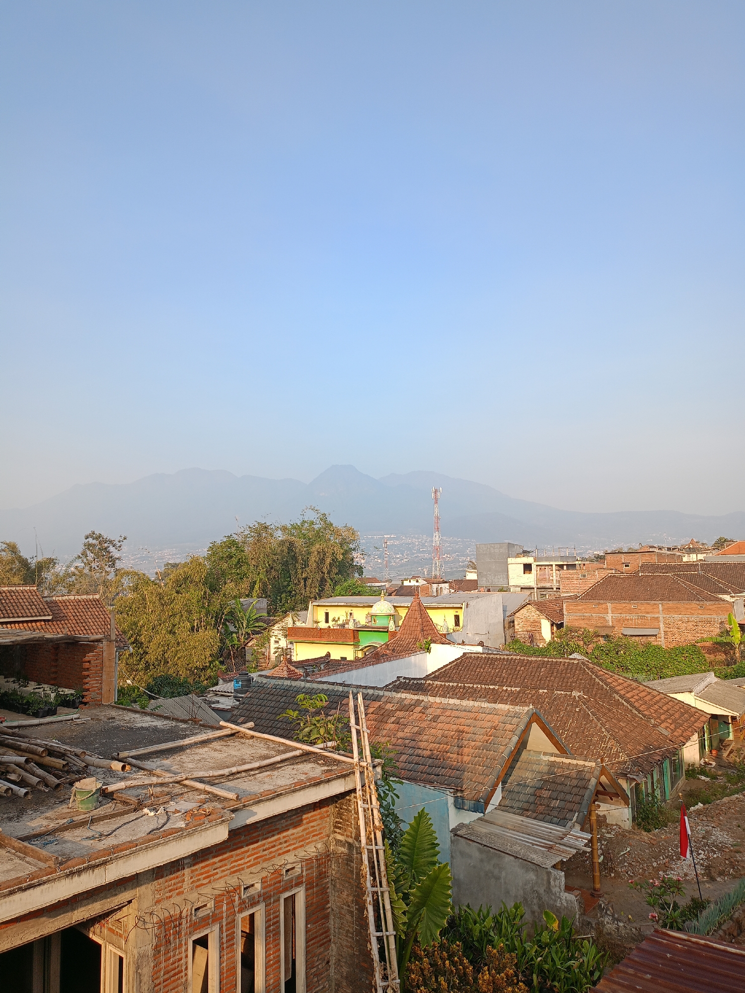 This is a landscape photograph taken from an elevated vantage point, likely a rooftop, overlooking a residential area in the city of Batu, Indonesia. The foreground is dominated by the rooftops of various houses, some under construction and others appearing lived-in. There are exposed brick walls, unfinished concrete structures, and bundles of bamboo poles and scaffolding, indicating ongoing building activity.
The middle ground features more houses with distinctive terracotta tile roofs, interspersed with lush green trees and vegetation, including bamboo groves. A yellow building with a green trim stands out among the residential structures. In the distance, a tall, white and red telecommunications tower is visible, a common sight in urban and semi-urban landscapes.
The background is characterized by rolling hills and mountains, shrouded in a light haze, characteristic of a clear but not perfectly crisp atmosphere. The sky is a pale blue, transitioning to a lighter shade near the horizon. The lighting suggests it is either early morning or late afternoon, with soft sunlight illuminating the scene and casting gentle shadows. There are no people visible in the frame. The overall impression is of a densely populated, somewhat rural or suburban setting within a mountainous region.