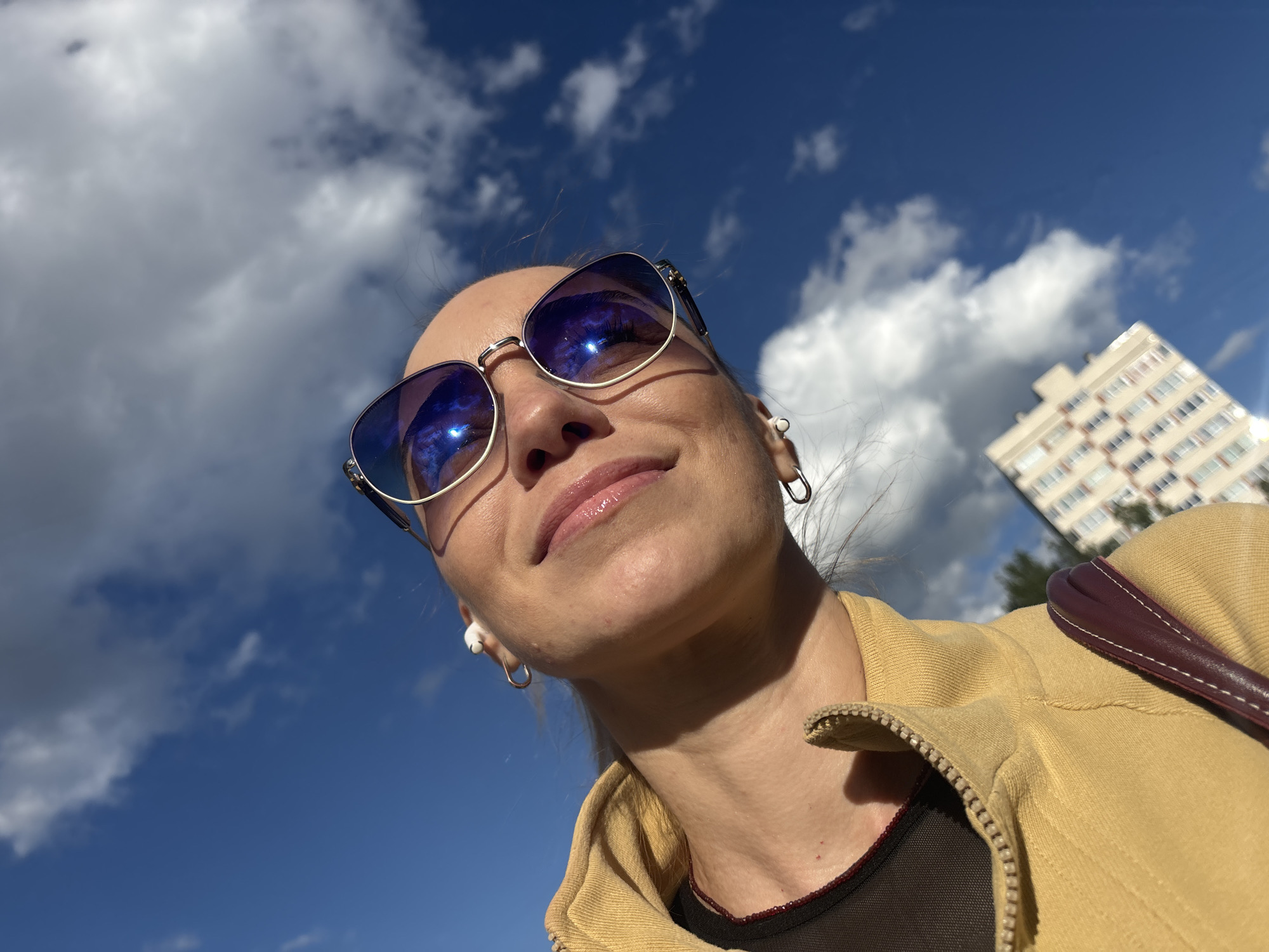 A close-up, low-angle, outdoor, selfie-style photograph captures a woman's face and upper torso against a bright blue sky with scattered white clouds. She is wearing large, metallic-rimmed sunglasses with blue lenses that reflect the sky and clouds. The woman has fair skin, light-colored lips, and is wearing white earbuds in her ears. She is also wearing a yellow hooded jacket, partially zipped, with a dark layer underneath. A portion of a dark red handbag strap is visible on her left shoulder. In the background, out of focus, is a light-colored building with many windows. The overall lighting suggests a sunny day. The image does not provide specific details about the location within a city, only that it is outdoors in Russia.