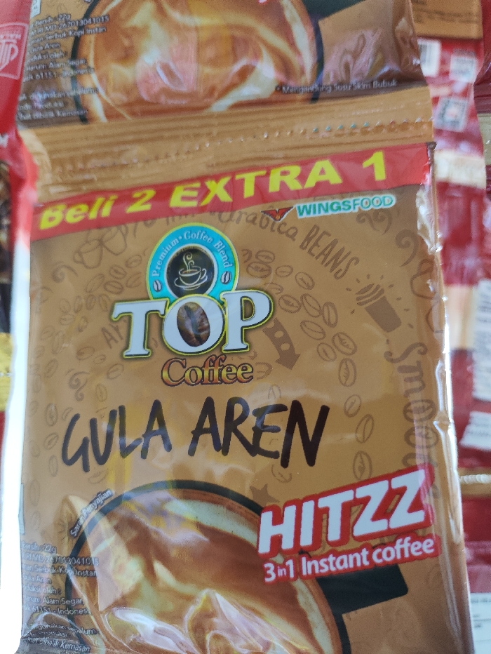 The image displays a close-up of a "TOP Coffee GULA AREN HITZZ" 3-in-1 instant coffee package, likely in a retail or home setting in Pubian, Indonesia. The packaging is a predominantly brown and gold sachet with a glossy finish, featuring the "TOP Coffee" brand logo in white and gold.

Key text visible on the package includes the product name "GULA AREN" (palm sugar) in a dark, handwritten-style font, and "HITZZ 3 in 1 Instant coffee" in a prominent white and red font. The manufacturer, "WINGSFOOD," is identified. A promotional offer, "Beli 2 EXTRA 1" (Buy 2 Get 1 Extra), is highlighted in yellow and red at the top. Other details include "Premium Coffee Blend" and "Arabica BEANS."

The package design incorporates illustrations of coffee beans scattered across the background, a stylized coffee cup icon, and a depiction of a prepared coffee drink, which appears to be a light brown, creamy beverage. Small print includes regulatory codes like "MD 267013041015" and "PIMDI2703041015," ingredient information such as "Serbuk kopi instan" (Instant coffee powder) and "Mangandung Soser Skim Bubuk" (Contains Skim Milk Powder), and serving instructions ("Saran Penyajian"). The phrase "Alam Segar" (Fresh Nature) and "Indonesia" are also partly visible.

The overall scene suggests an indoor environment with bright, even lighting. No individuals are present in the image. The product appears to be one of many, with hints of other packages visible in the background, implying a stack or display. The image is taken at a slight downward angle, focusing on the front of the coffee sachet.