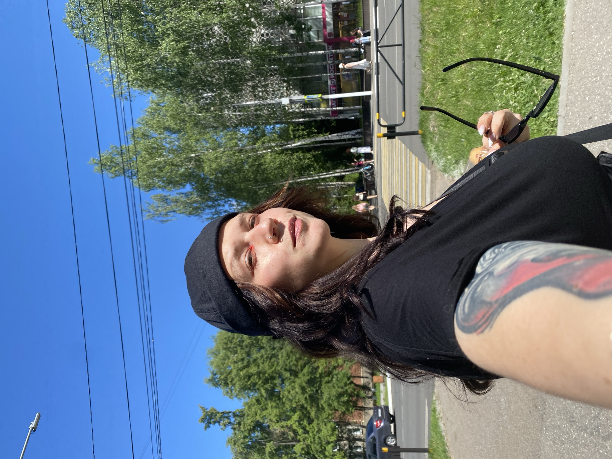 An outdoor, eye-level, medium shot captures a young woman in a black beanie and black t-shirt, facing right, as she takes a selfie on a sidewalk in Chaikovsky, Russia. She has dark hair, nose jewelry, and red eyeshadow. Her left arm, with a prominent tattoo, is extended towards the camera, holding a small pastry. In her right hand, she holds a pair of black sunglasses. The background reveals a pedestrian crossing, a street with a few cars, a grassy area, and a row of birch trees lining the sidewalk in front of buildings, one with a sign that reads "КОСМЕТИК." The sky is clear blue, and there are power lines running overhead.