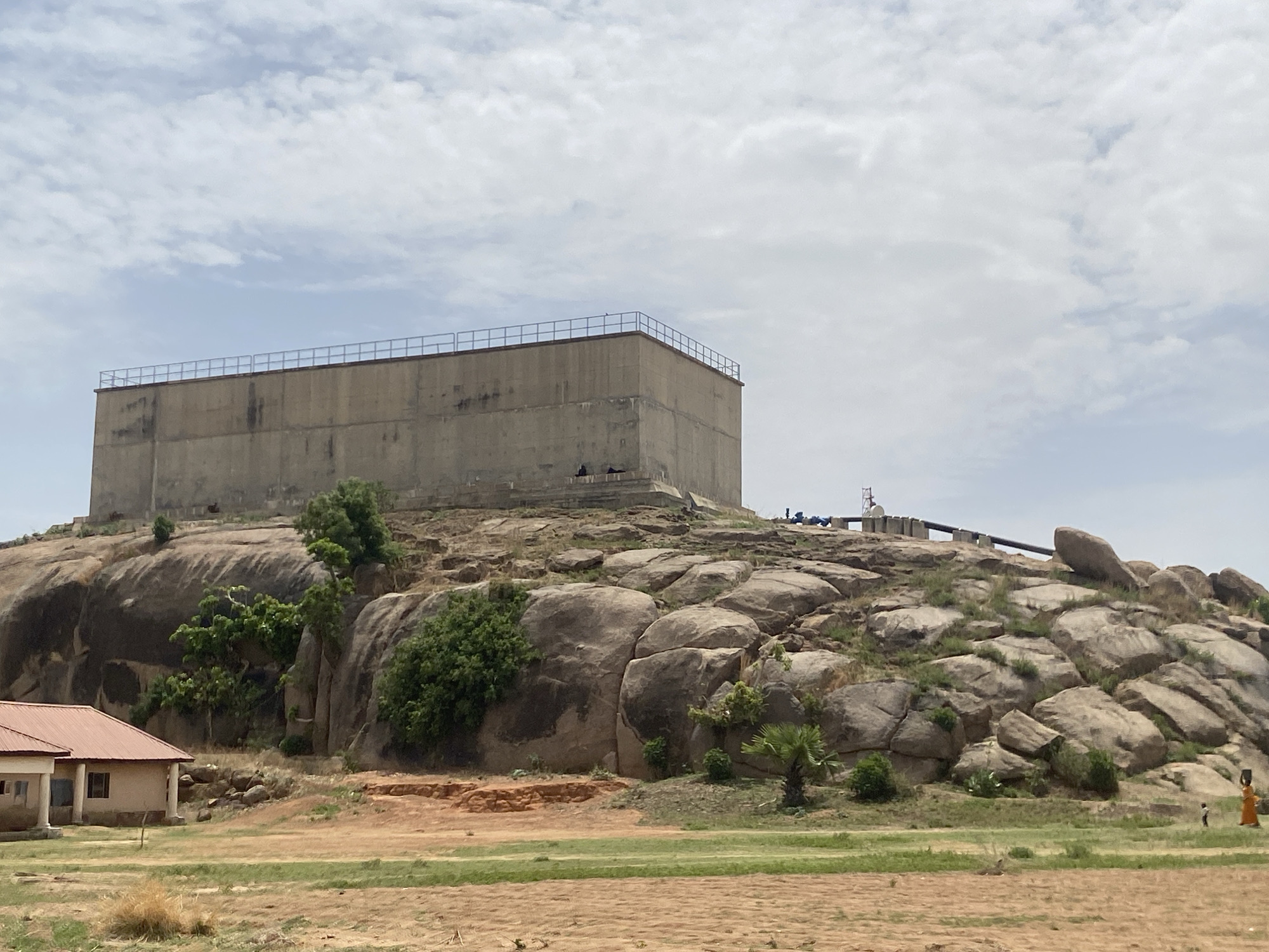 The image shows a large, rectangular, concrete reservoir structure atop a rocky hill in Kufena, Nigeria. The reservoir has a metal railing around its top perimeter. A pipeline runs along the rocky ground behind the reservoir. The hillside is covered with large, rounded rocks and sparse vegetation, including trees and palm-like plants. At the base of the hill is a small building with a red roof. The foreground consists of dry, sandy ground interspersed with patches of green grass. In the lower right corner, two figures appear to be walking, one wearing an orange robe. The sky is partly cloudy.