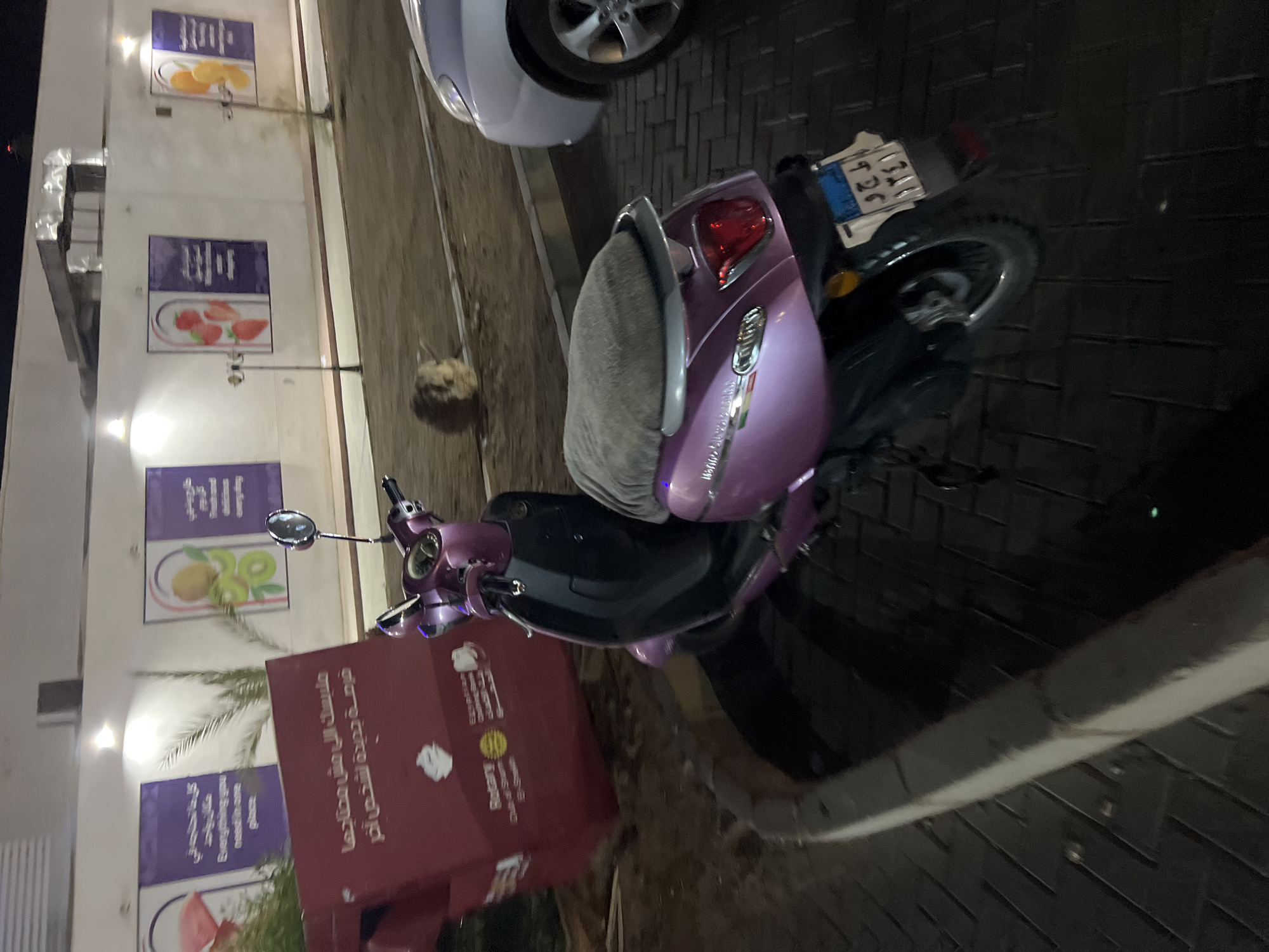 A parked pink and black scooter is shown outdoors at night in Sharm El Sheikh, Egypt. The scooter has a grey seat cover and is parked on a brick-like paved surface. Behind the scooter, a silver car is partially visible. In the background, there is a white building with several large, purple-tinted posters displaying images of fruit and text in Arabic and English. A large red bin or dumpster is also visible on the left side of the image, with text that appears to say "Rotary" and Arabic writing. A small, fuzzy object, possibly a rock or discarded item, is on the ground between the scooter and the building. The scene appears to be a street or public area in the city.