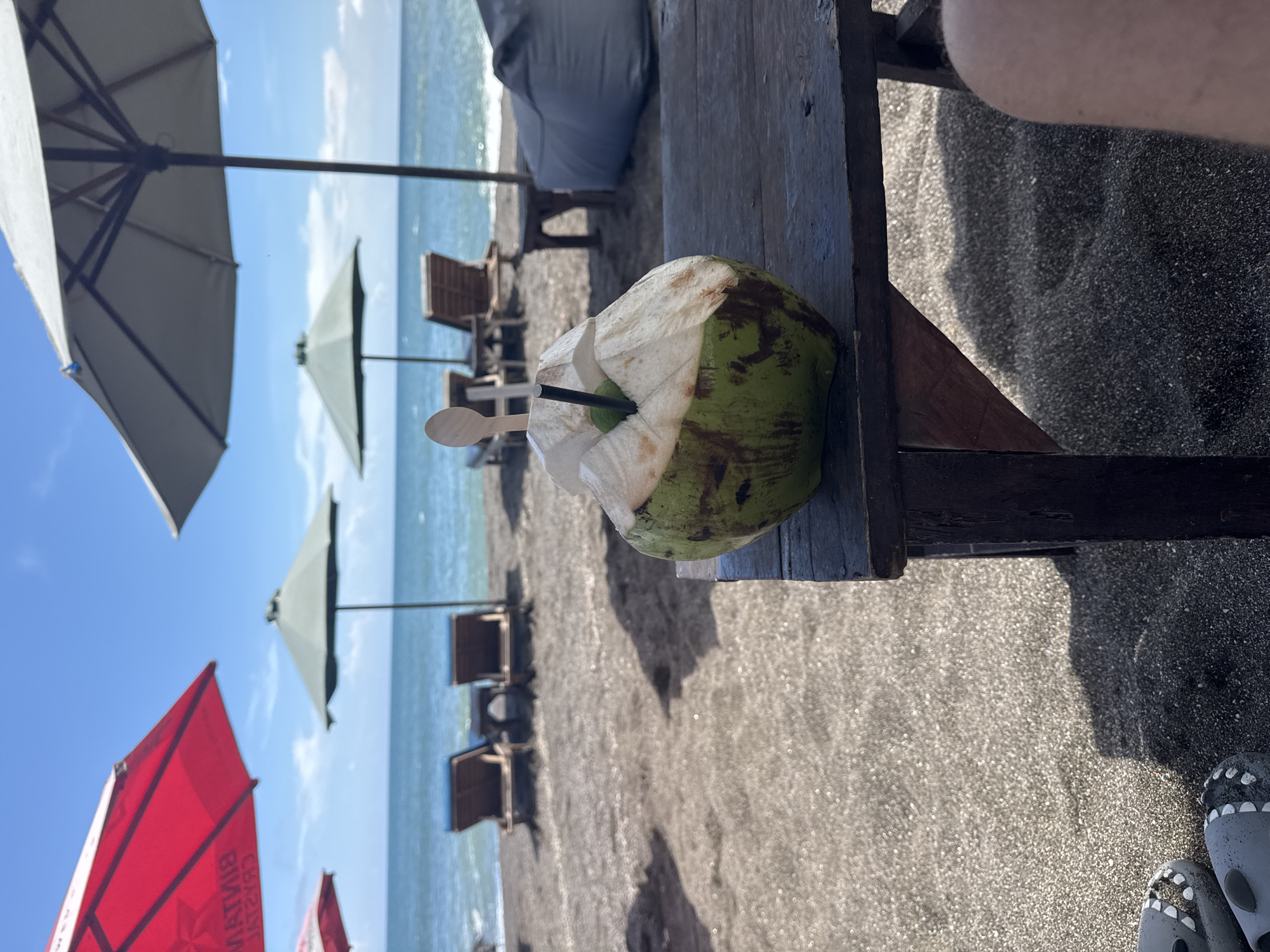 This image was taken on a beach in Canggu, Indonesia. The scene shows a young coconut with a straw and spoon in it sitting on a wooden table on the beach. There are several beach umbrellas and lounge chairs behind the table, with the ocean visible in the background. A person is standing next to the table with their feet and shoes in the lower right corner. The beach has gray sand. The weather appears to be sunny.