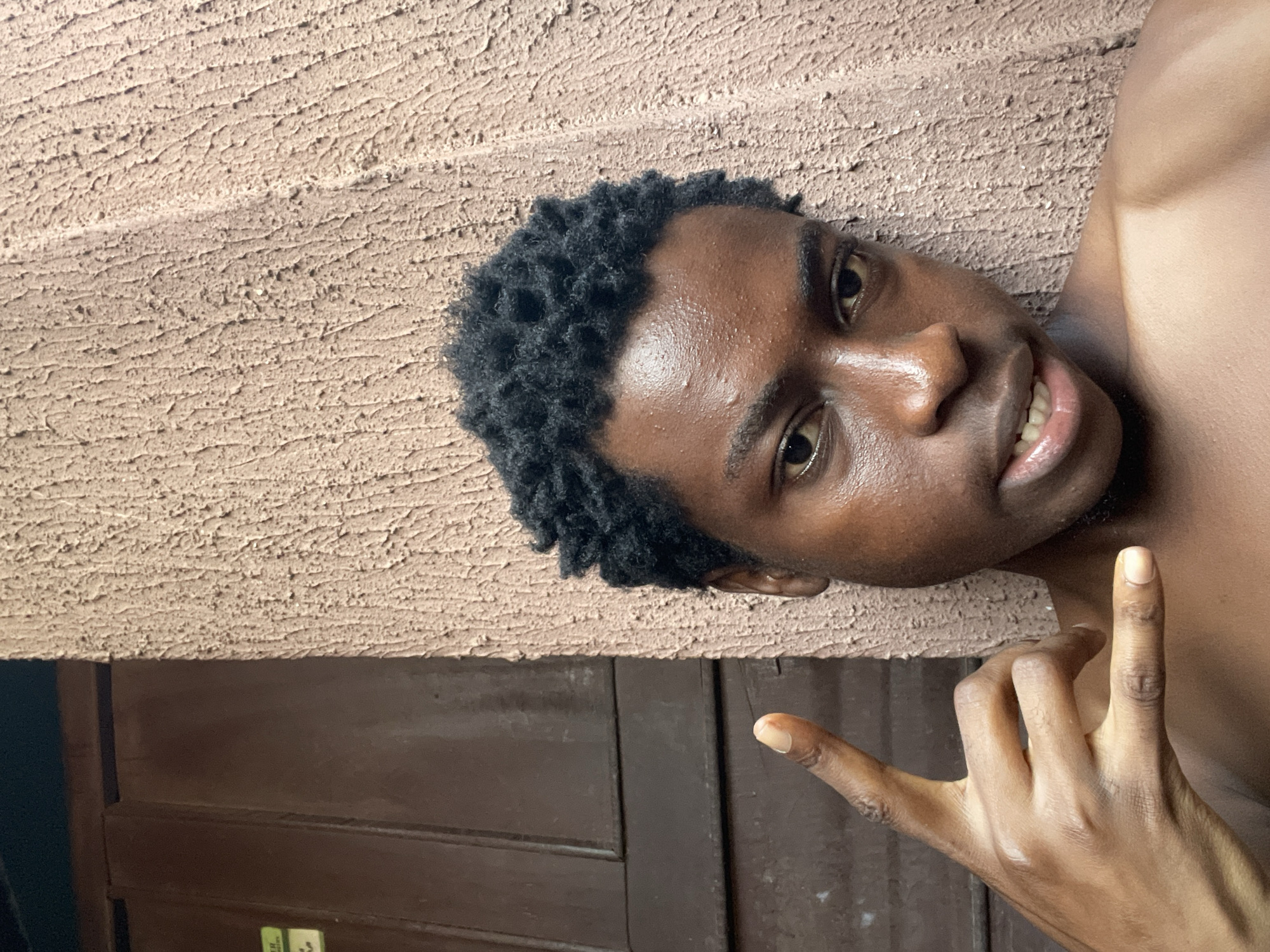 This media file features a close-up, low-angle shot of a young Black man lying down and looking at the camera, situated against a textured, tan-colored wall. His head is tilted, and his dark, curly hair is visible. He has a slight smile on his face, revealing his teeth, and his eyes are open, looking directly forward. He is wearing no shirt, exposing his upper body. His right hand is raised, with his index and pinky fingers extended, forming a "rock on" gesture. The background behind him is split between the textured wall and dark brown wooden doors.
The scene appears to be outdoors, possibly in a residential area given the presence of doors. The lighting suggests it might be daytime, with natural light illuminating the man's face and skin. The overall mood seems relaxed and casual, with the subject appearing comfortable and playful. There is no visible text in the image. The location context provided is Nkpor, Nigeria, which suggests the setting is likely a warm climate.