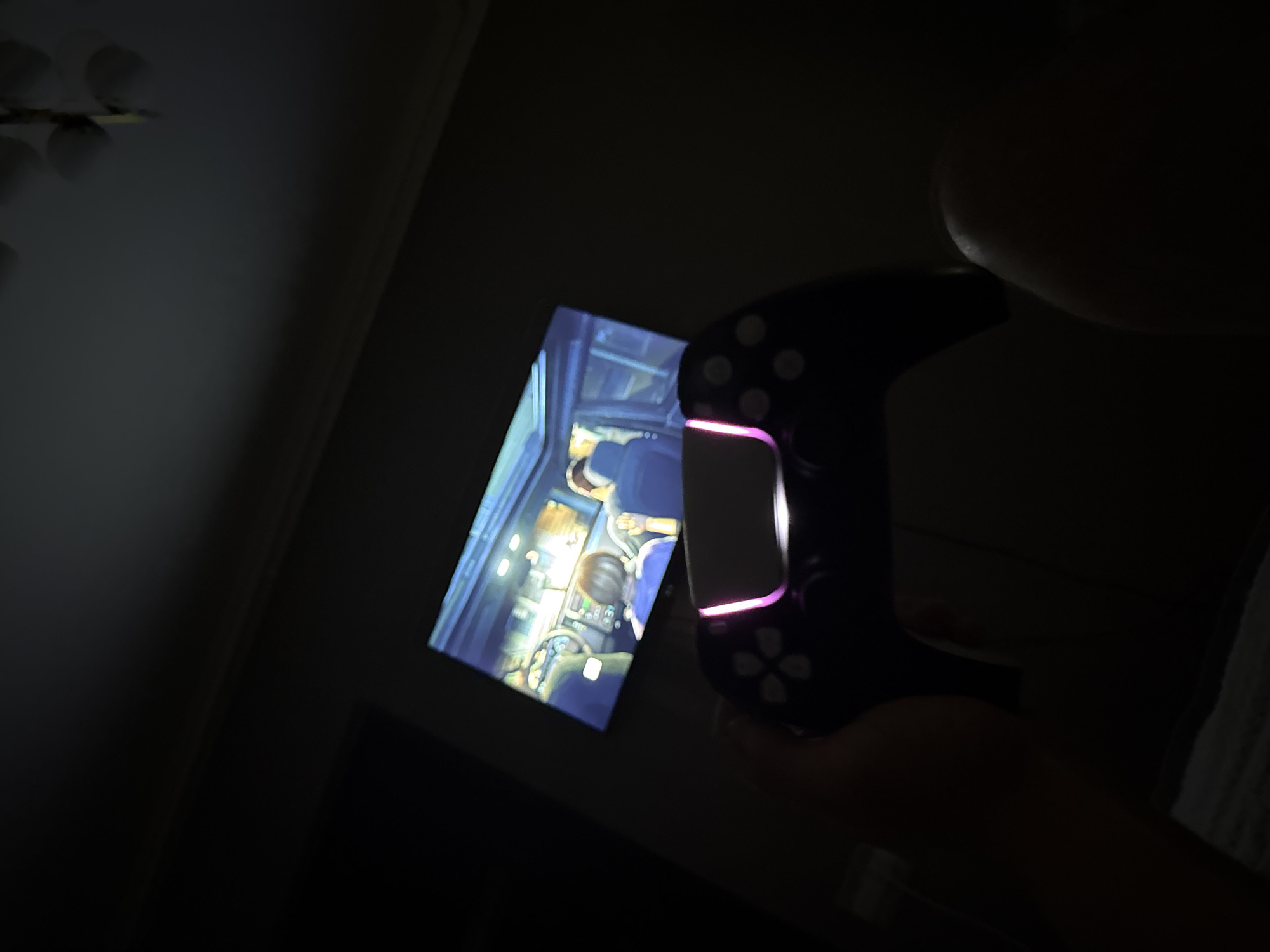 In a dimly lit room, a person's hand holds a black video game controller with glowing pink and white lights. The controller is positioned in the foreground, angled towards the viewer. In the background, a television screen displays a scene from a video game, showing the interior of a vehicle with animated characters. The overall ambiance suggests a private setting, likely a living room or bedroom, where someone is engaged in playing video games. The specific location in Tbilisi, Georgia, cannot be determined from the provided image.