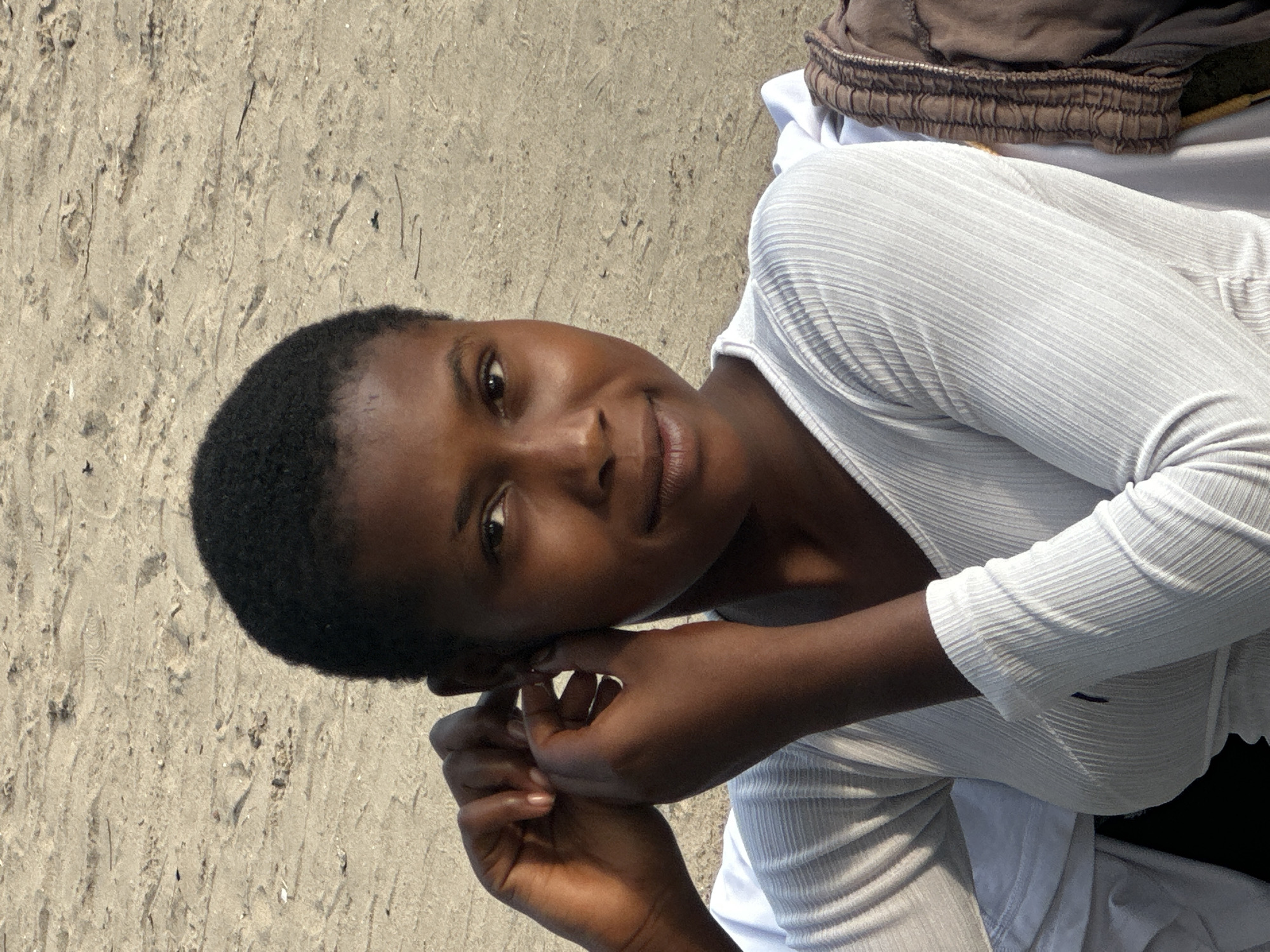 In Badagry, Nigeria, a young Black woman with short, dark hair is photographed outdoors, seated on a sandy surface. She is wearing a white, long-sleeved, ribbed top with a V-neck. Her right hand is raised to her left ear, appearing to adjust an earring or her ear. Her gaze is directed towards the camera, and she has a subtle smile. The background consists of sandy ground, suggesting a beach or a sandy area. Clothing, possibly a brown garment, is visible to the right of the frame, resting behind the woman.